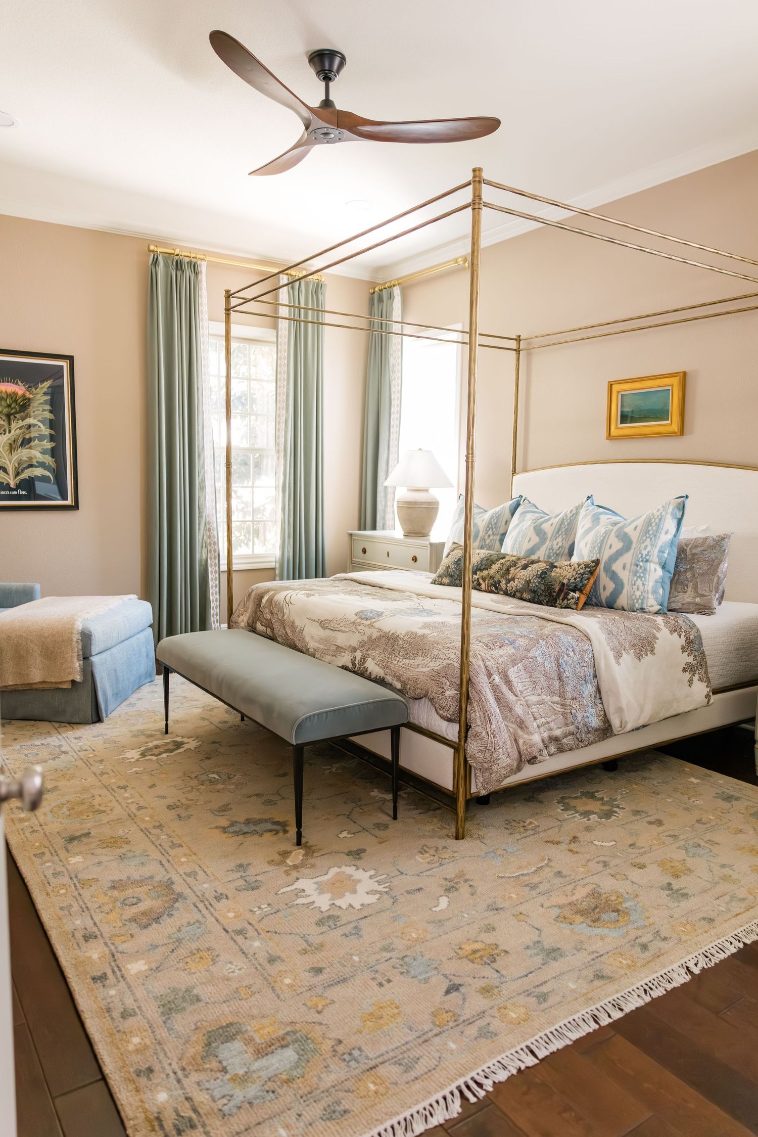 Serene bedroom with brass canopy bed, blue curtains, floral rug, ceiling fan, and layered decorative pillows.