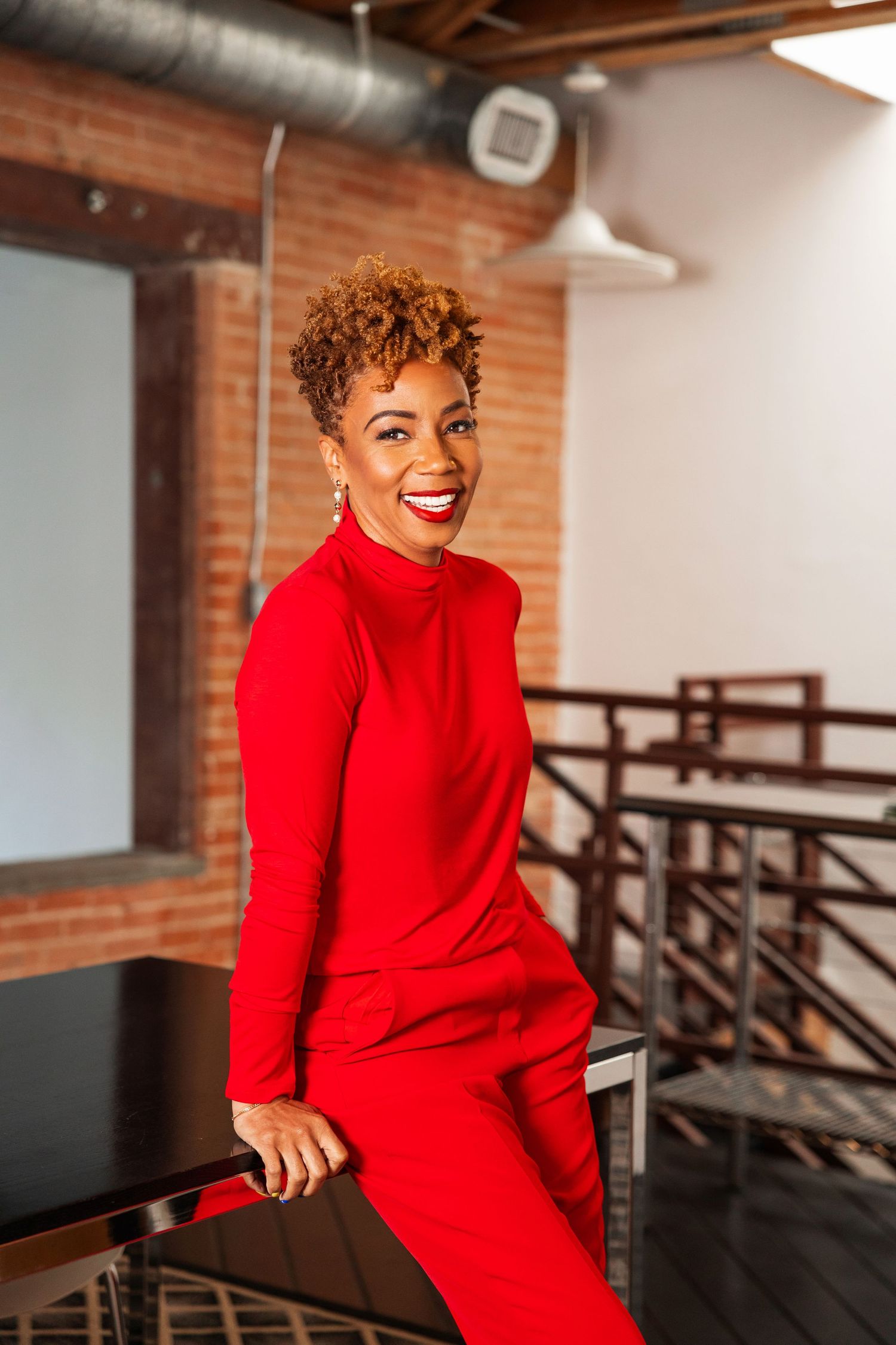 Smiling woman in red turtleneck jumpsuit leaning on railing in brick-walled loft space.
