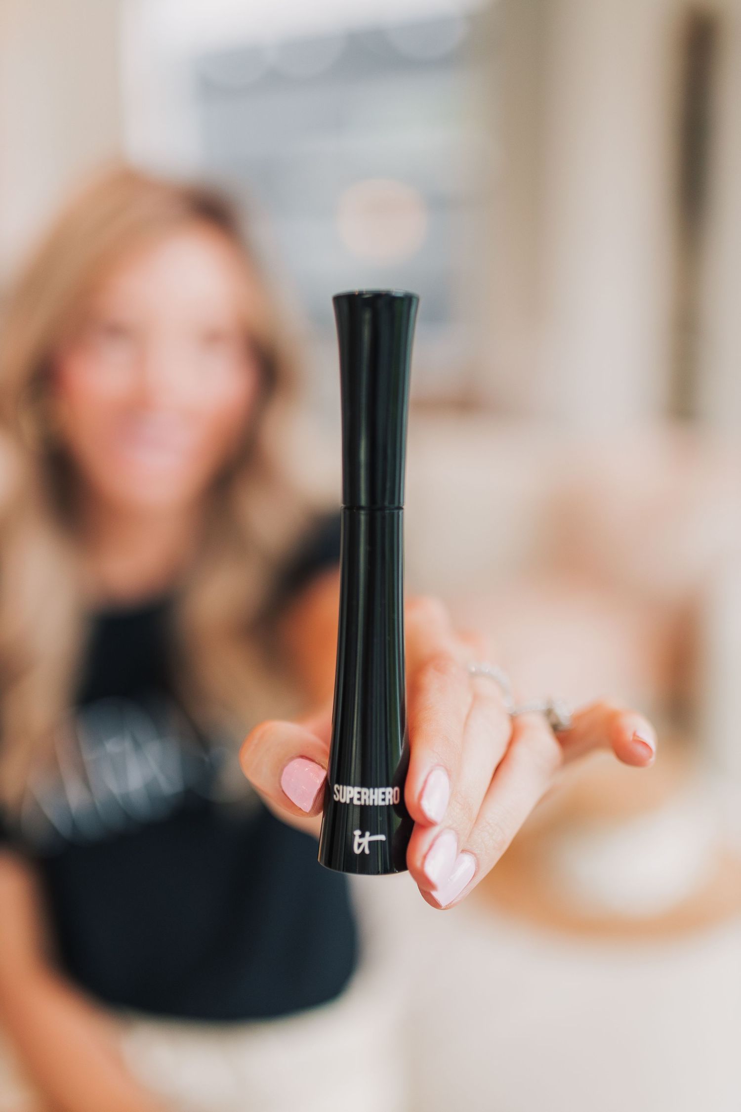 Hand holding IT Cosmetics Superhero mascara tube close to camera with blurred background.