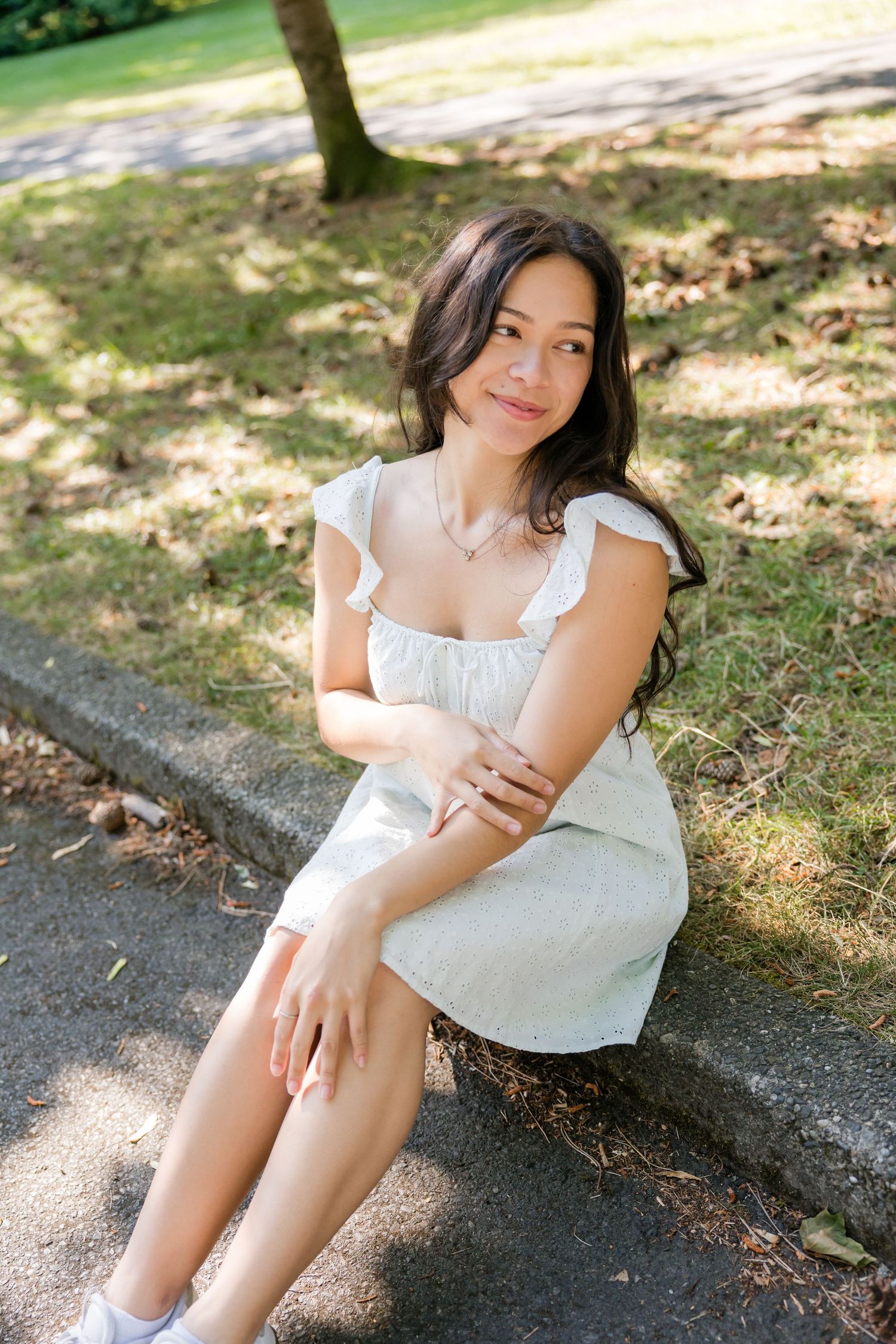 Young woman in white sundress sits on a park curb, smiling softly in dappled afternoon sunlight.