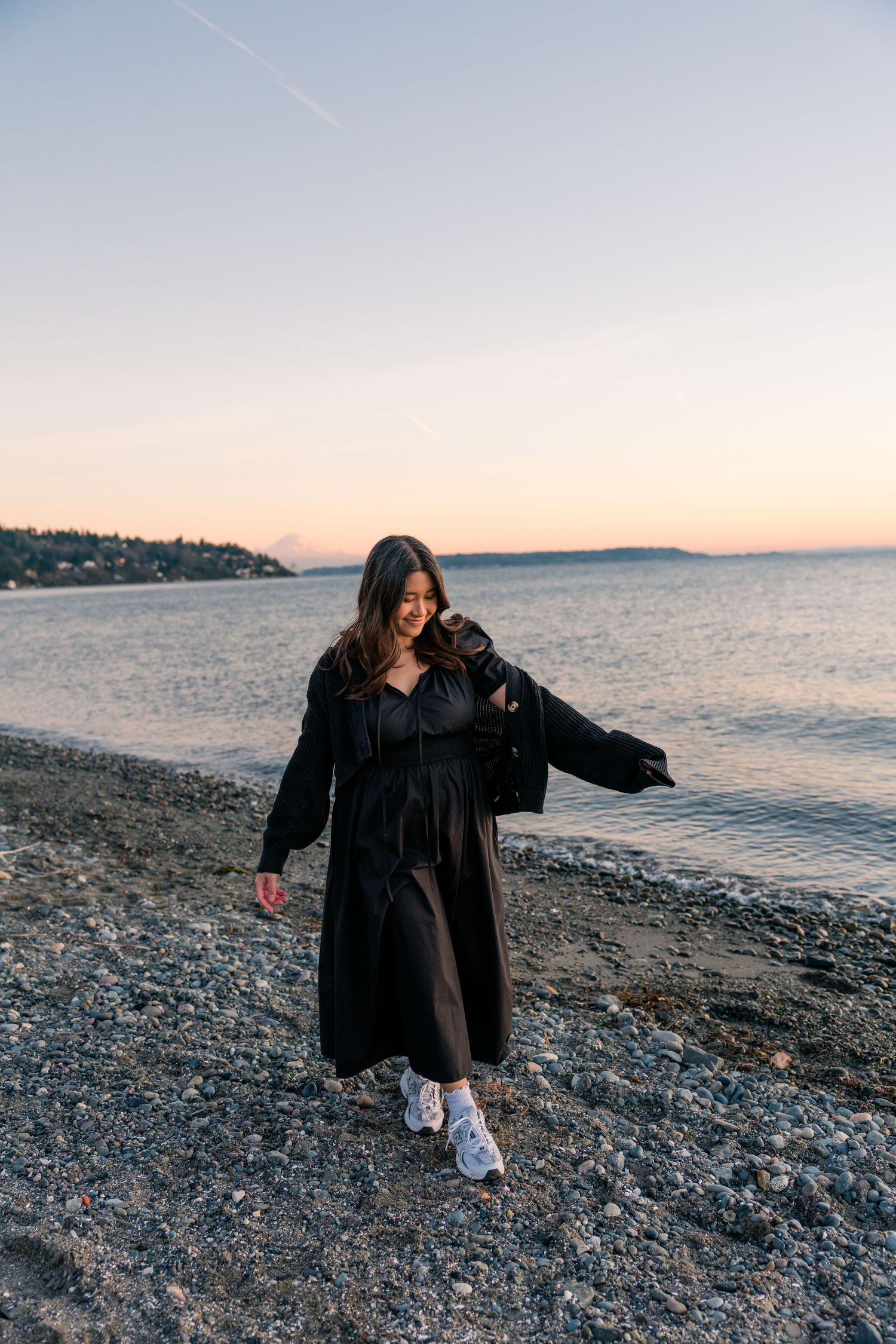 Cheerful woman in black midi dress and white sneakers strolls along a pebble beach at golden hour.