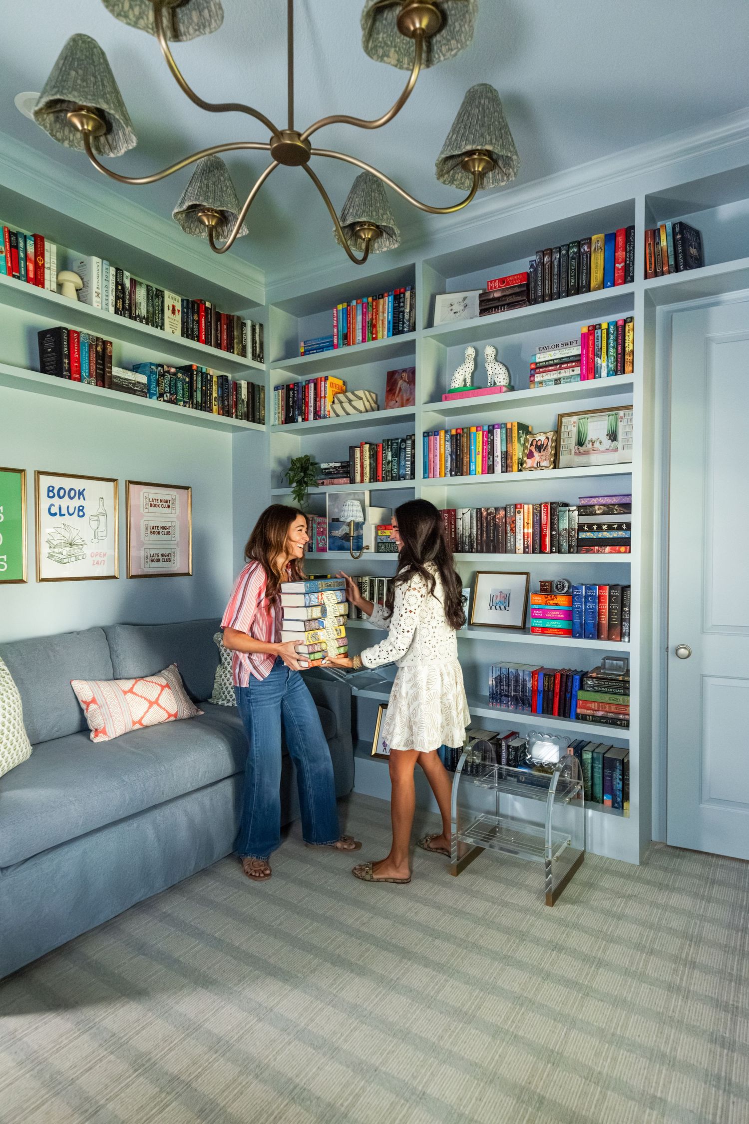Light blue home library with floor-to-ceiling bookshelves, gray sofa, brass chandelier, and two women organizing books.