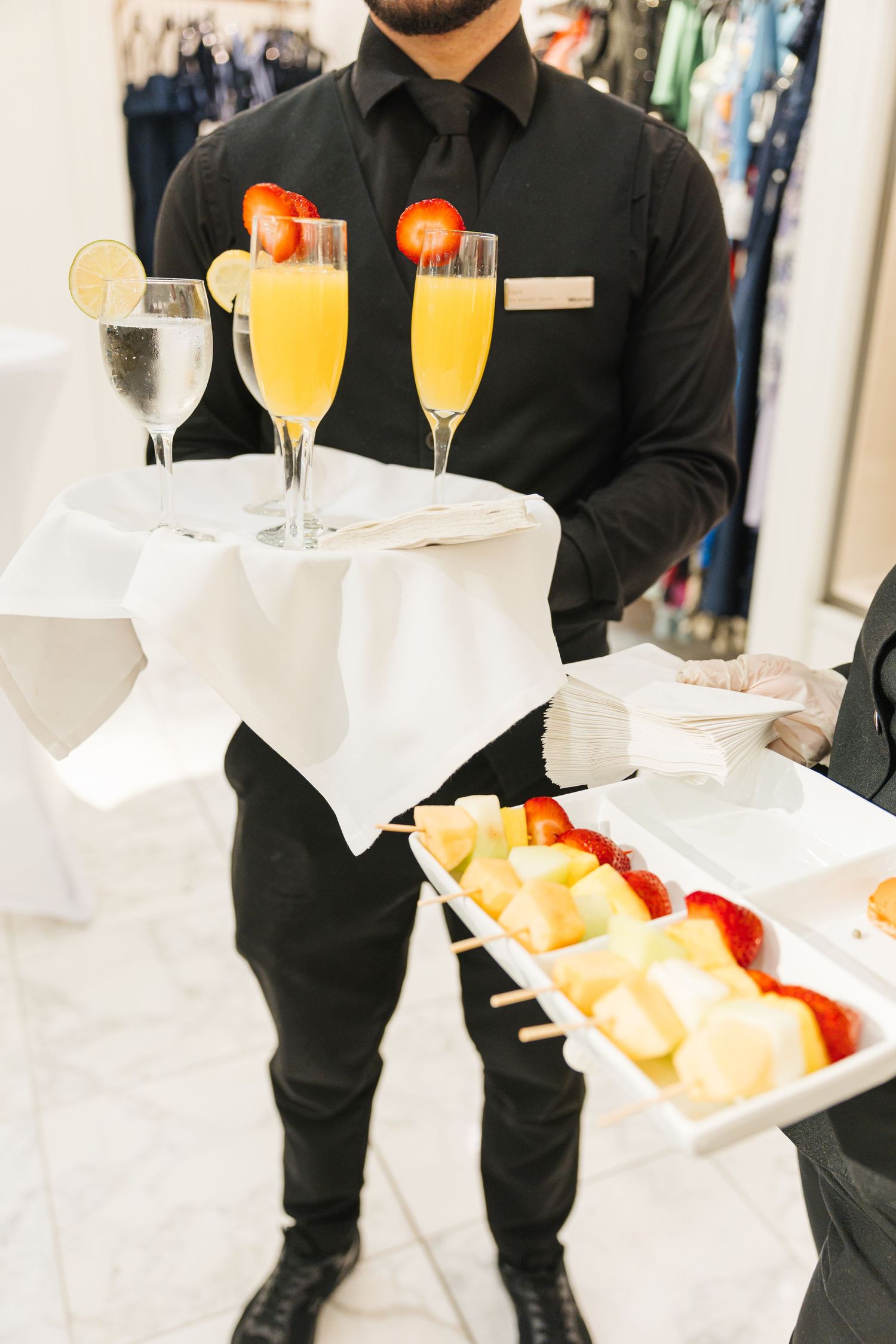 Waiter in black uniform serving mimosas and fresh fruit skewers on white trays at event.