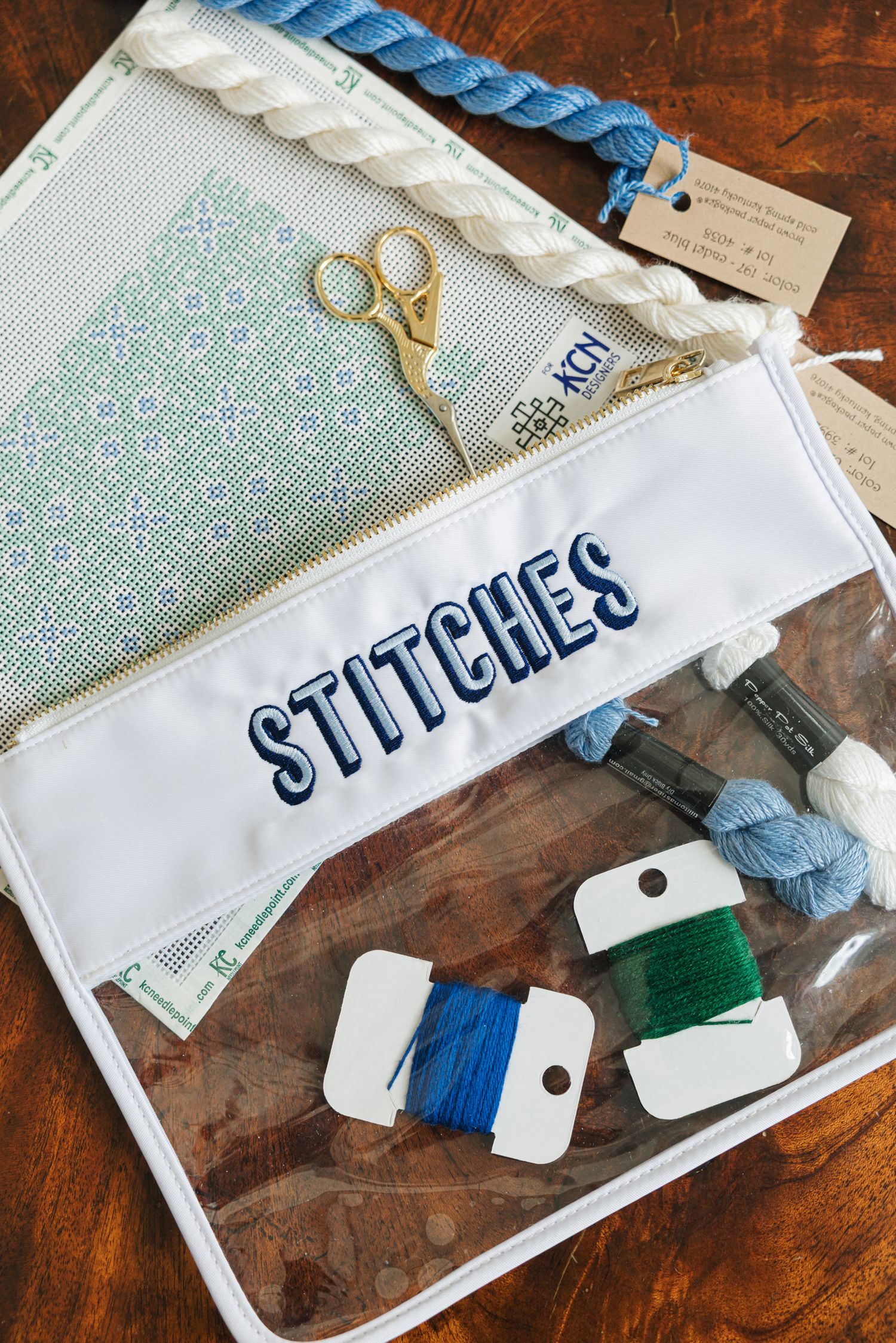 Needlepoint craft supplies with scissors, thread, and clear pouch labeled STITCHES on wood surface.