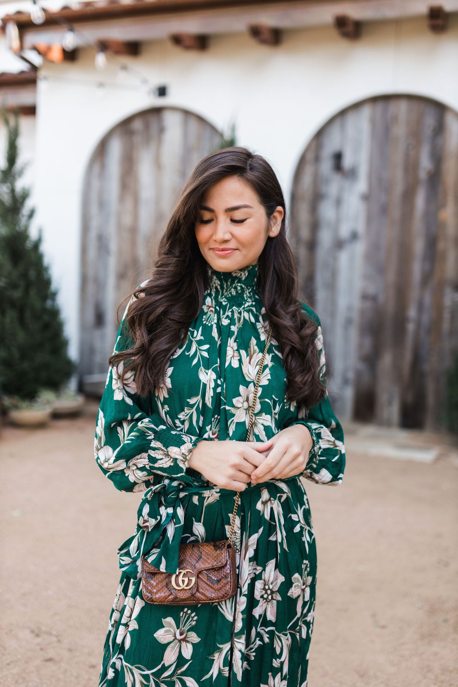 Woman in green floral maxi dress with brown Gucci bag poses outdoors near rustic wooden doors.
