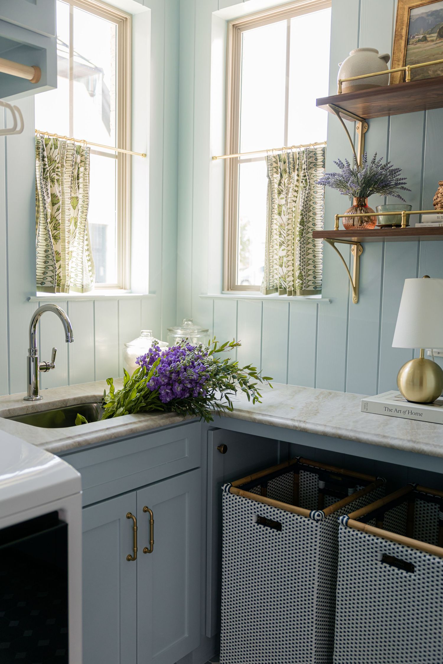 Light blue laundry room with marble countertop, farmhouse sink, purple flowers, woven baskets, and patterned curtains.