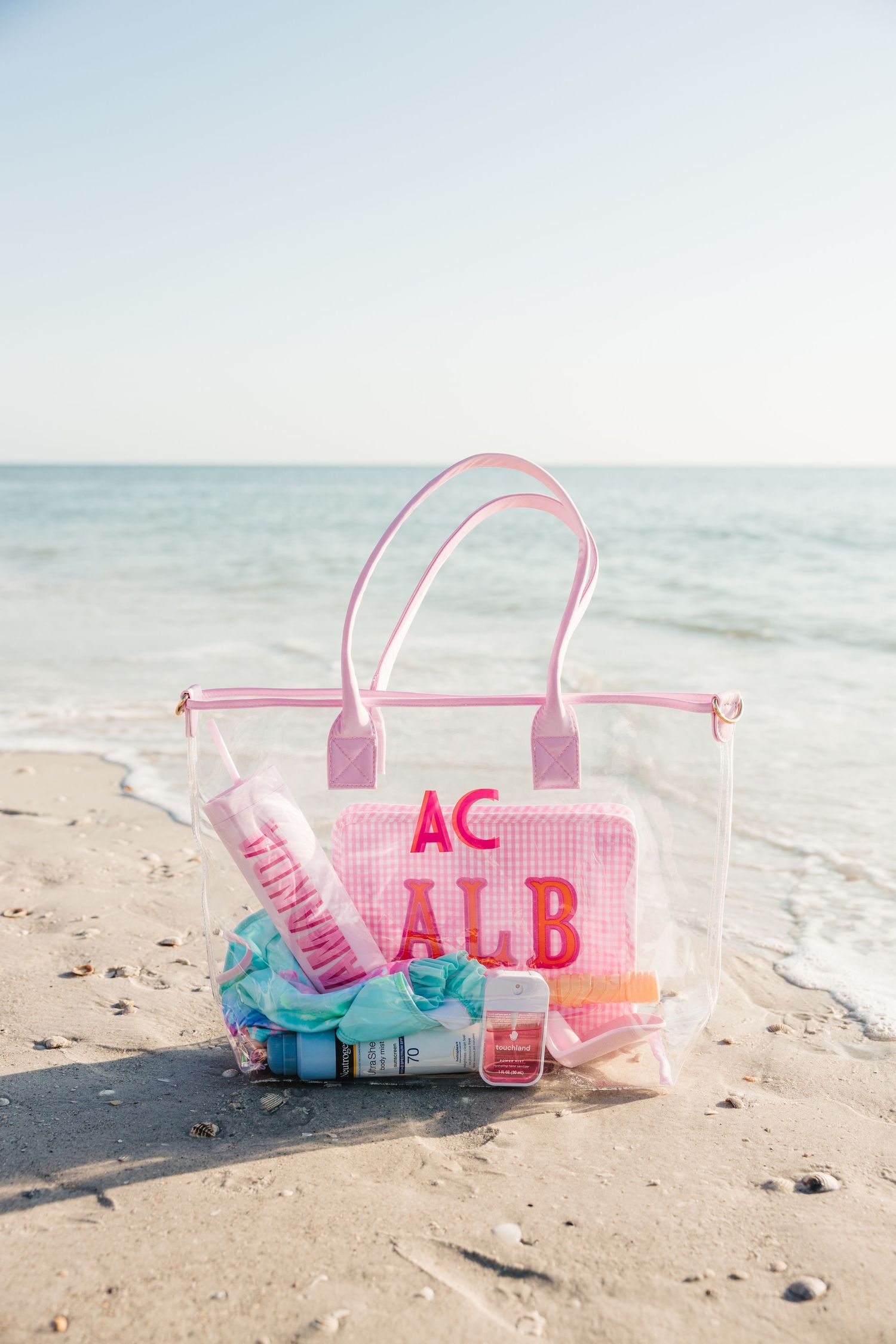 Pink monogrammed clear beach tote bag filled with sunscreen and accessories sitting on sandy beach.