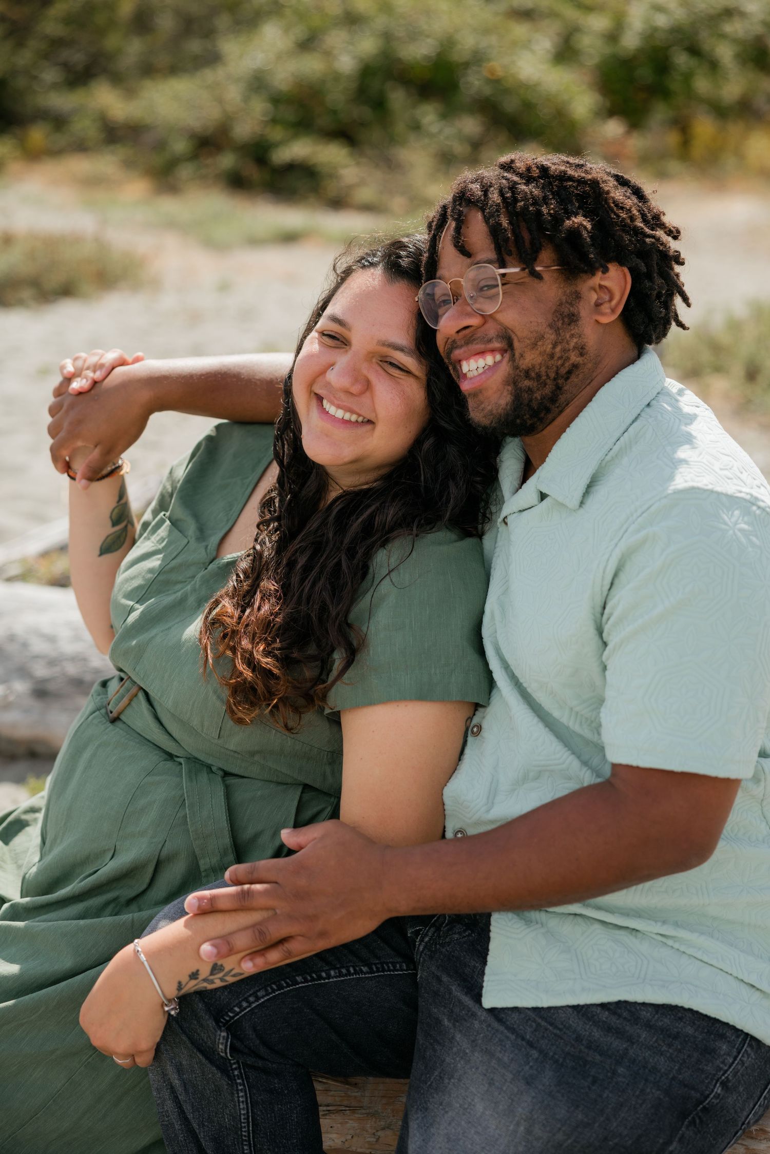 Smiling interracial couple embraces closely while sitting outdoors in a sunny natural setting.