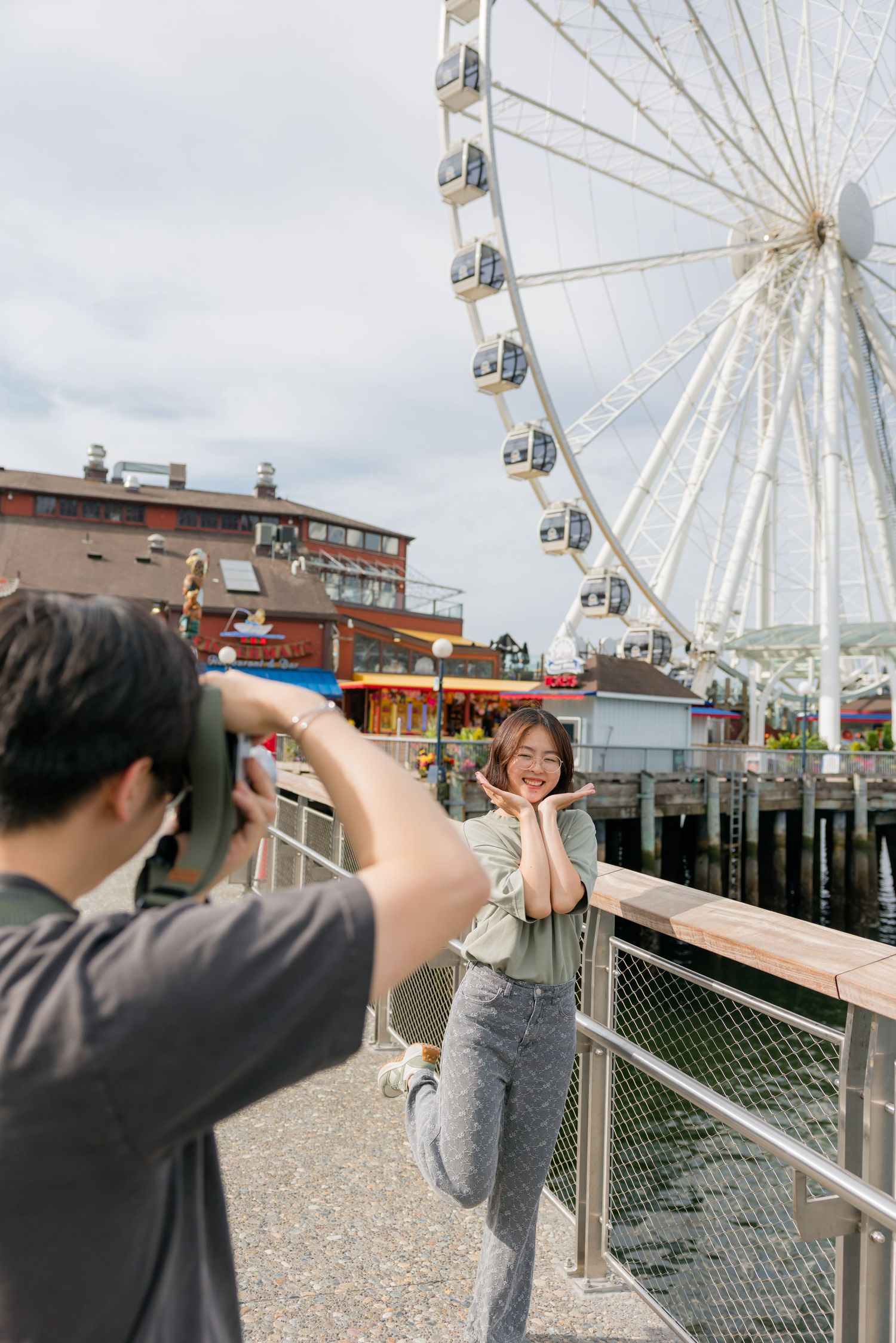 Young woman posing happily while man photographs her near Seattle's Great Wheel ferris wheel.