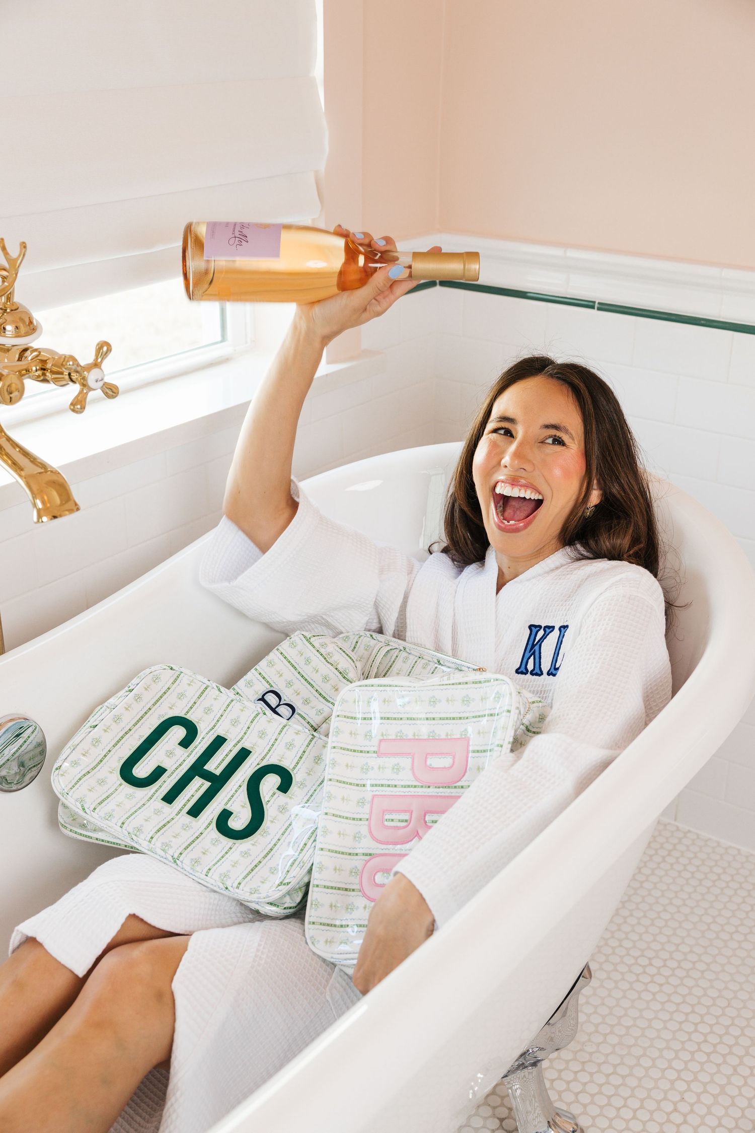 A cheerful woman in a white monogram robe holds a rose wine bottle while sitting in a clawfoot bathtub.