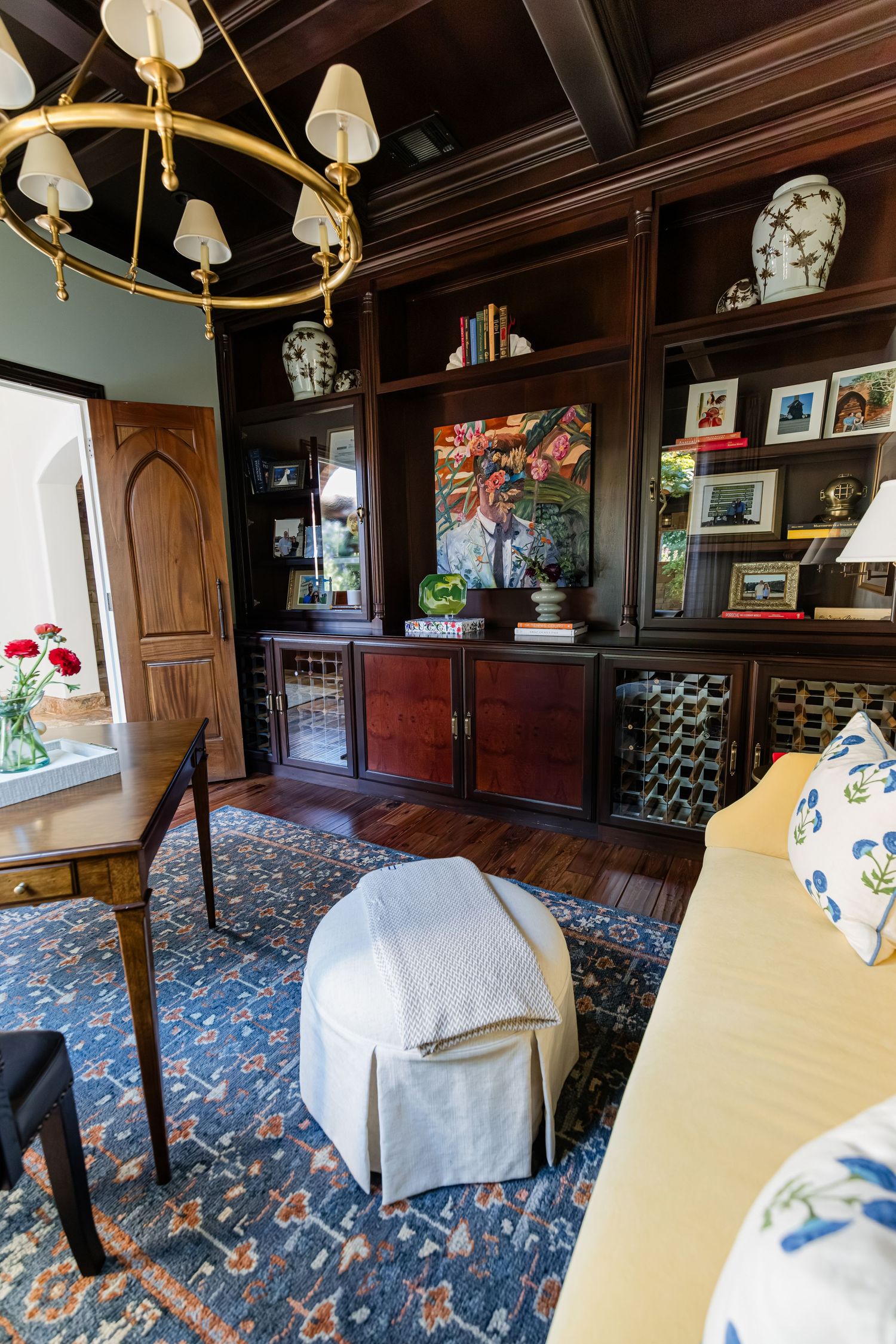Dark wood built-in shelving with brass chandelier, blue patterned rug, and yellow sofa in sophisticated study.