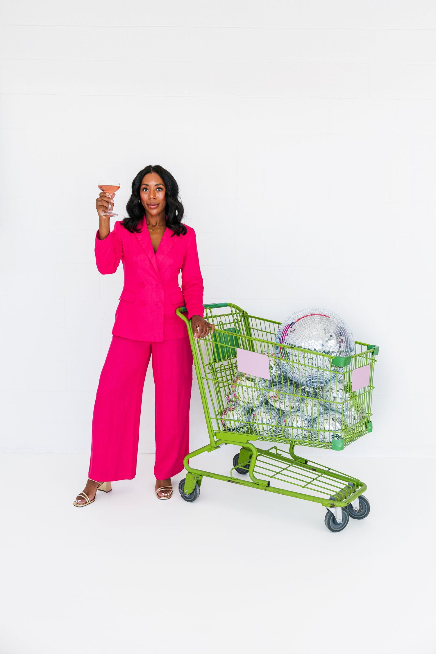 Woman in bold pink pantsuit holding cocktail beside green shopping cart filled with disco balls on white background.