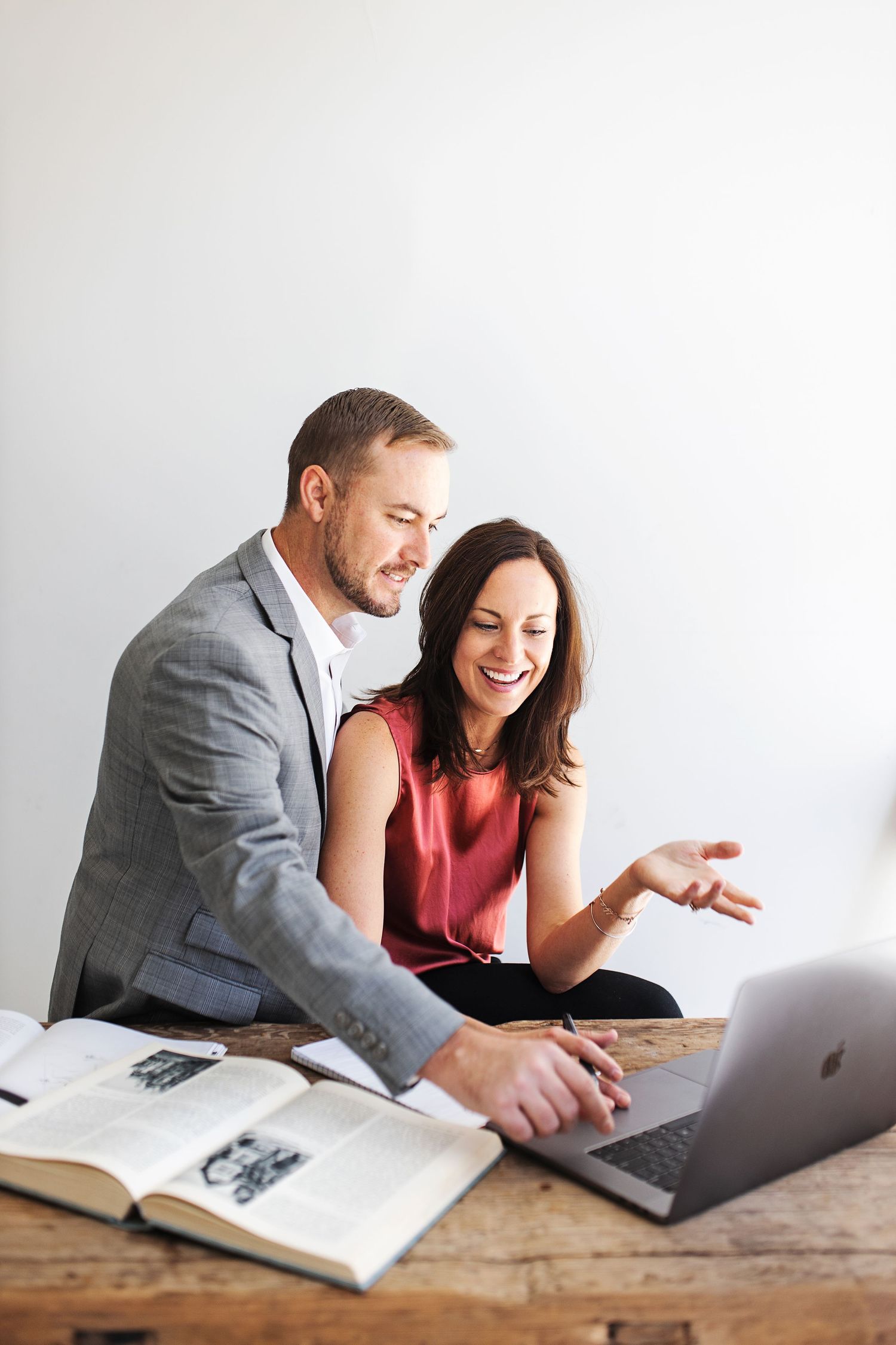 A man in a gray blazer and woman in a red top laugh together while viewing a laptop on a wooden desk.