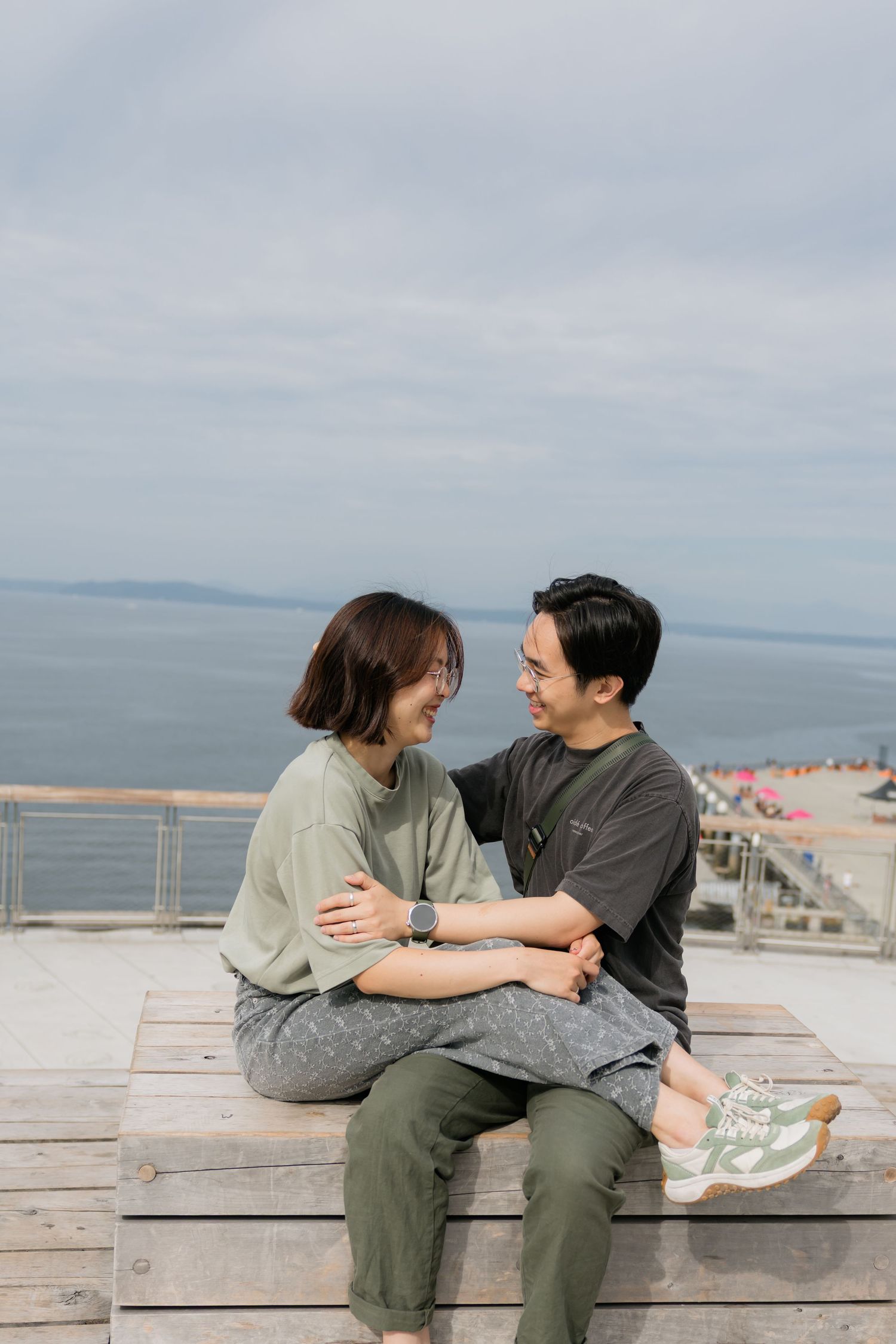 A couple sits together on a wooden deck overlooking the ocean, smiling and holding hands on a cloudy day.