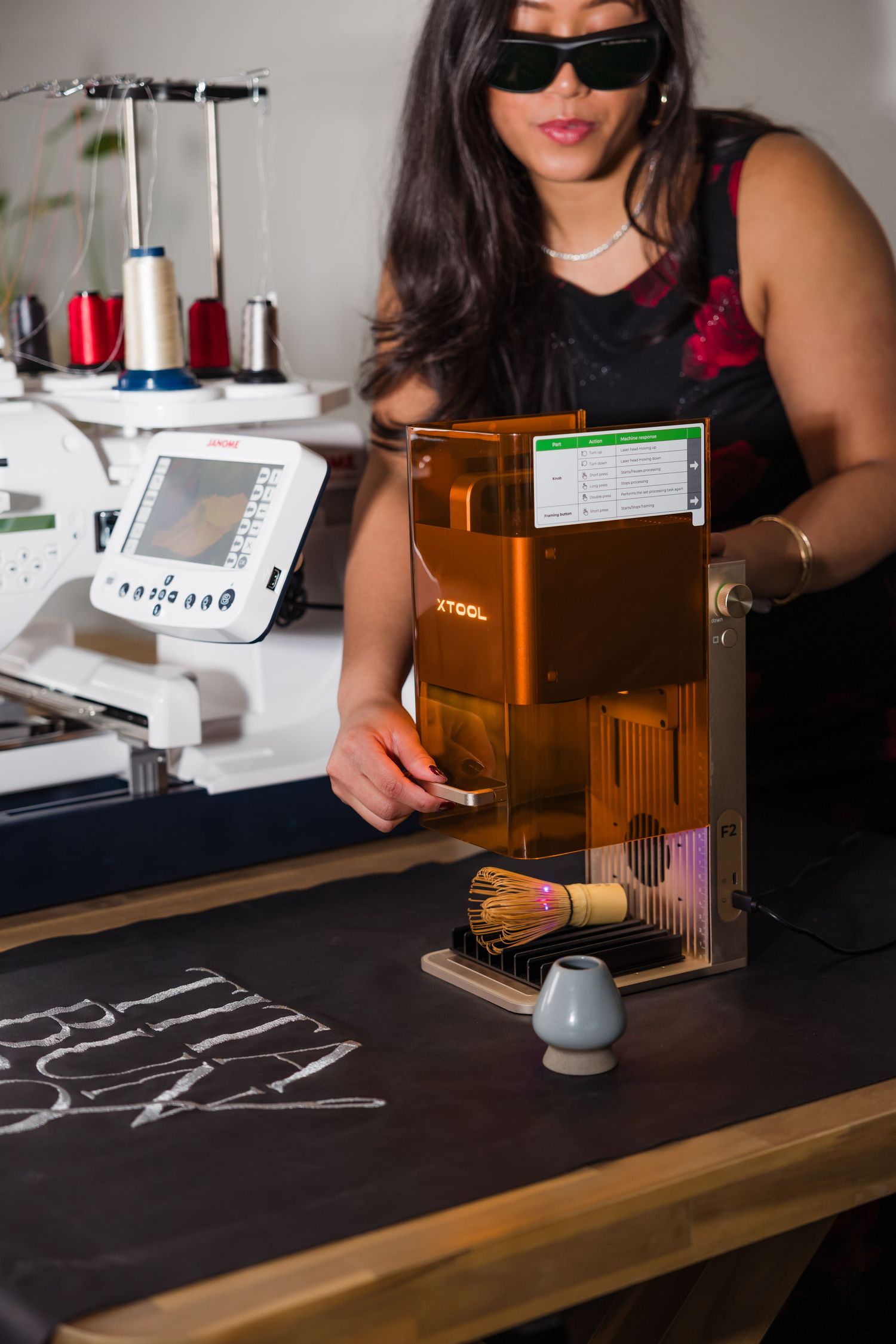 Woman in sunglasses operating an xTool laser engraver on a dark fabric near an embroidery machine in a craft studio.