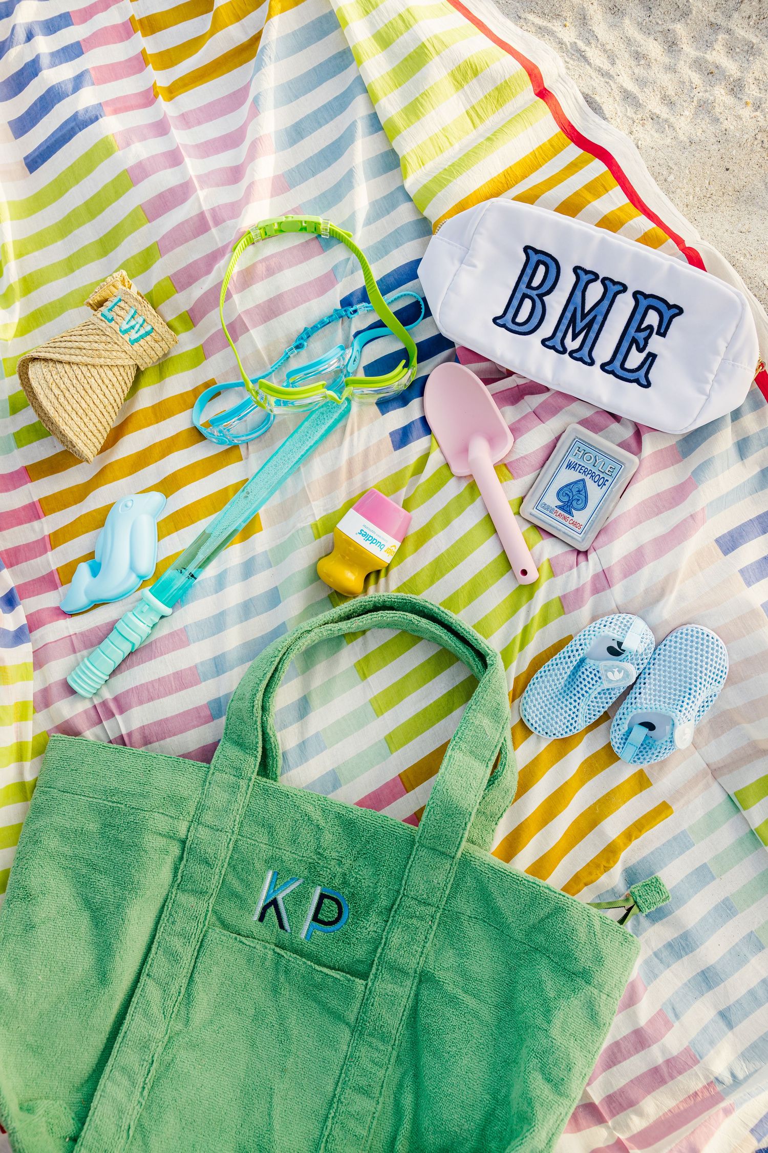 Colorful monogrammed green tote bag with beach toys, goggles, and accessories on a striped towel.