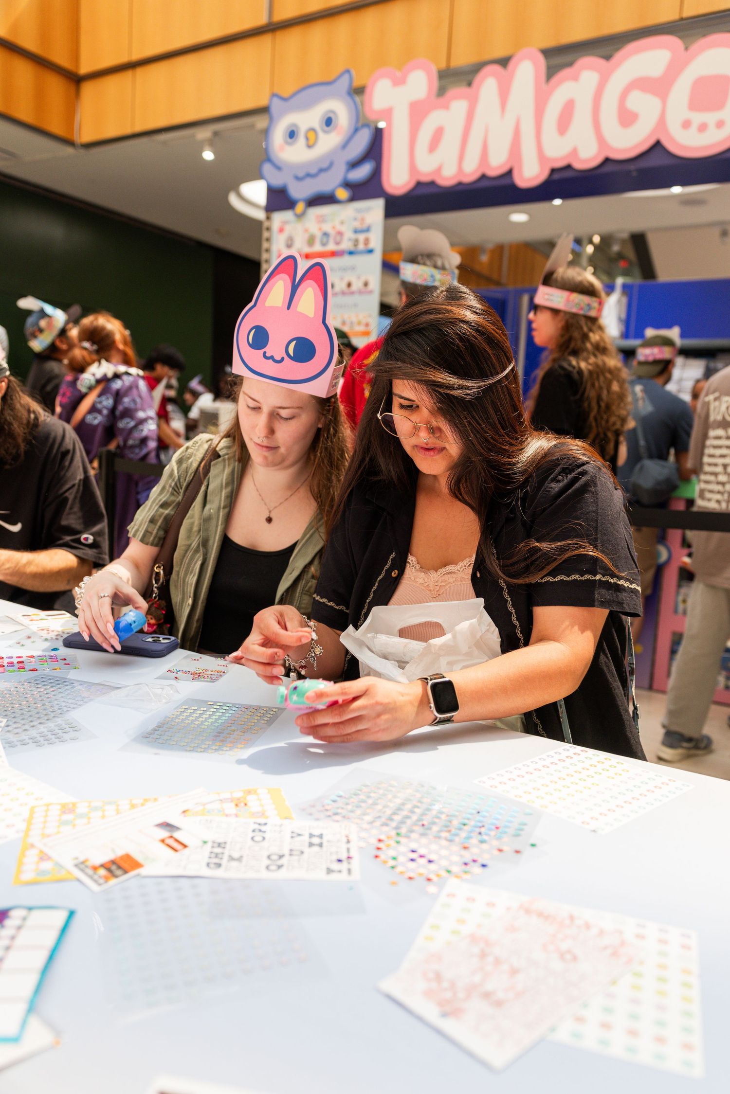 Two women craft at a Tamagotchi-themed activity table inside a shopping mall store.