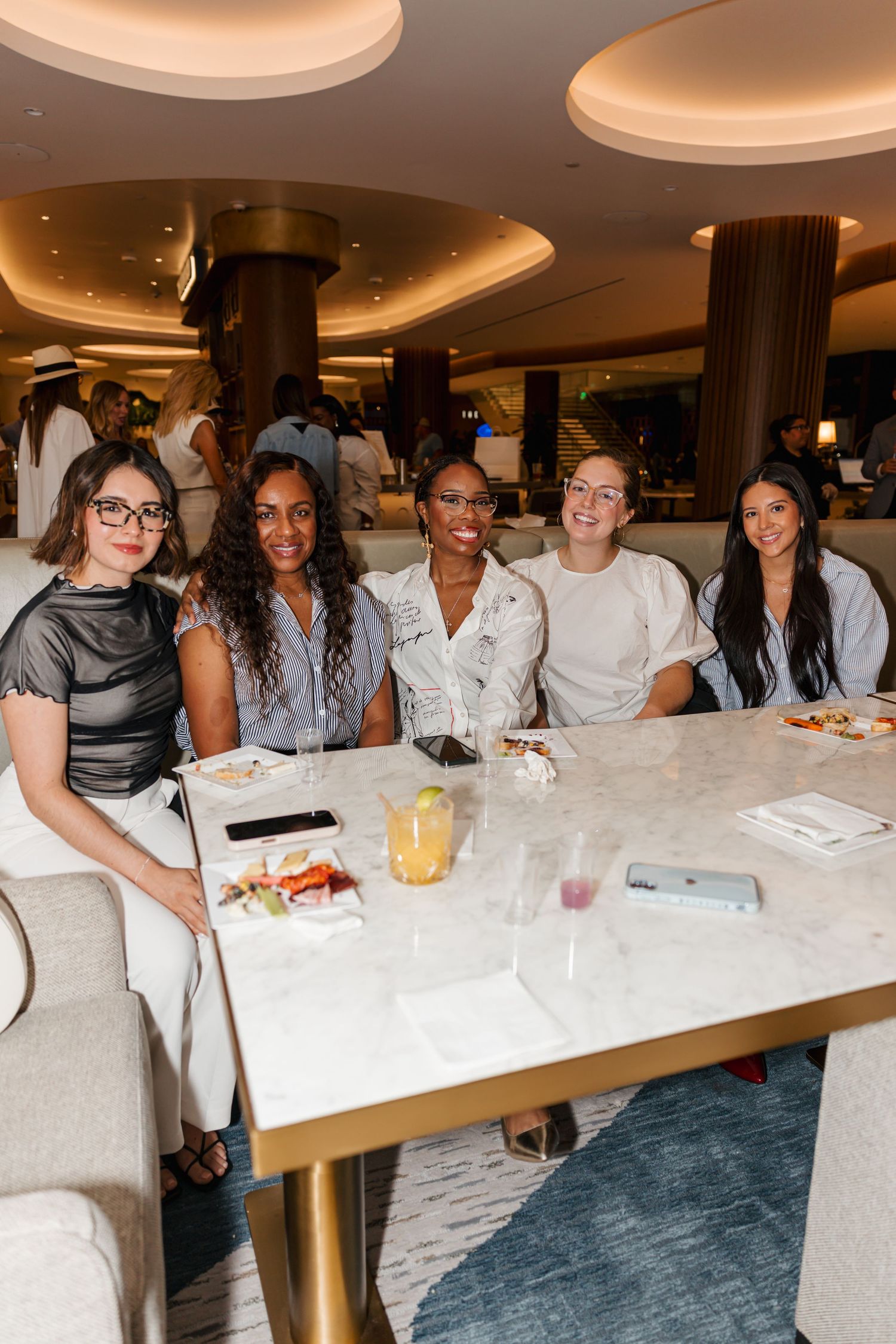 Five smiling women seated together at a marble dining table in an upscale restaurant.
