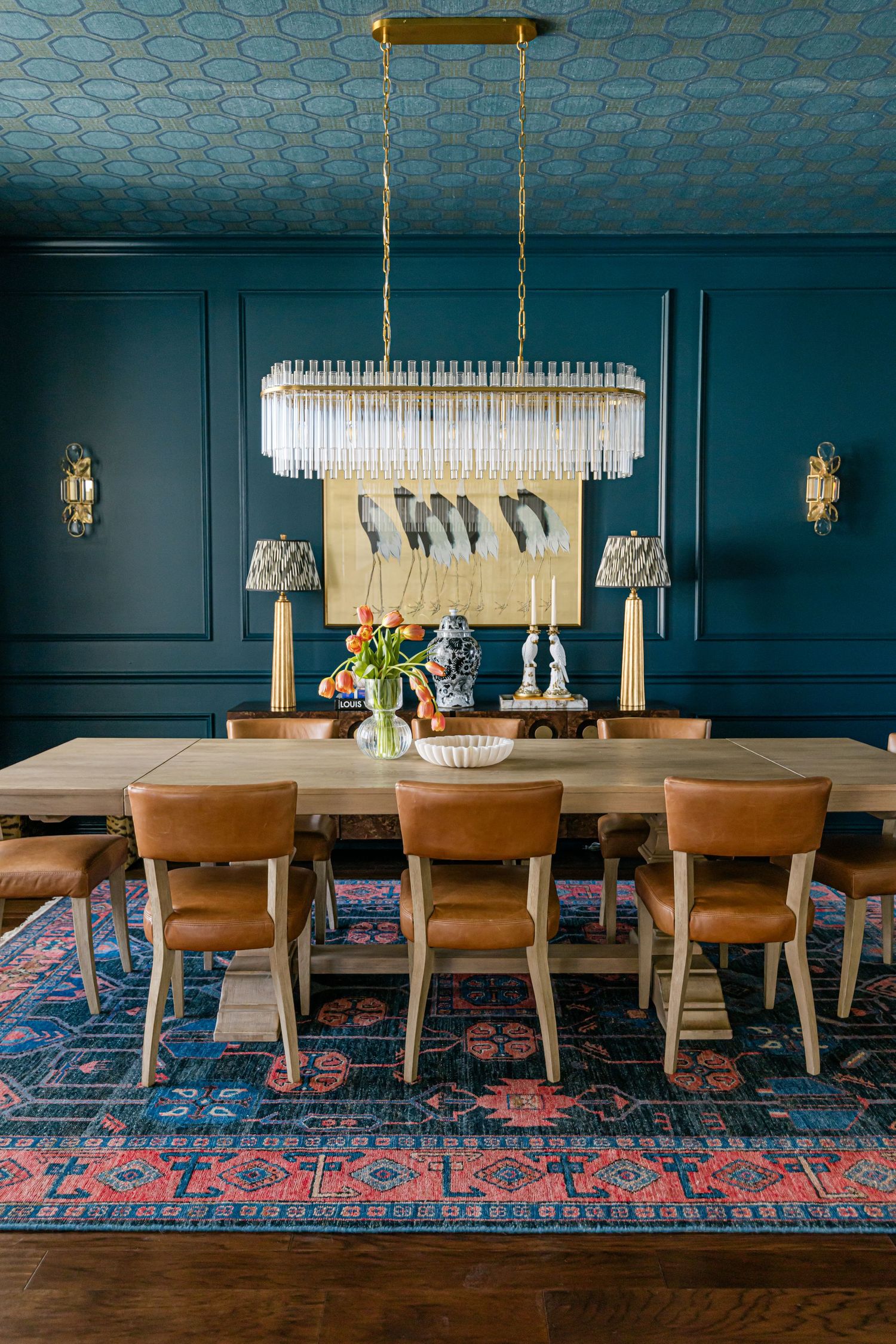 Elegant teal dining room with crystal chandelier, leather chairs, wood table, patterned rug, and decorative bird artwork.