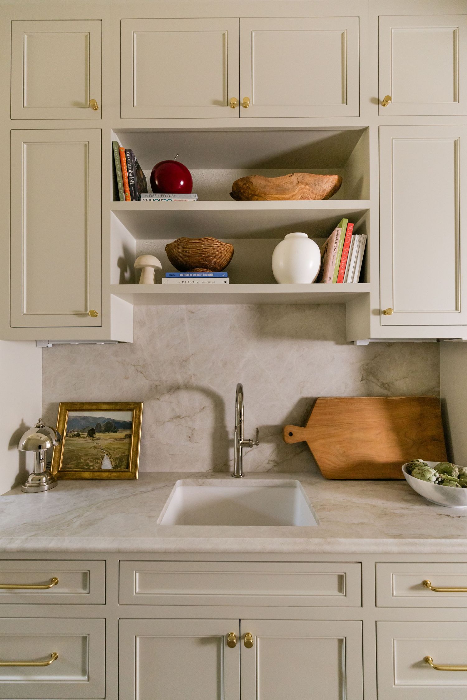 Cream kitchen cabinets with brass pulls, marble backsplash, open shelving with decor, and undermount sink.