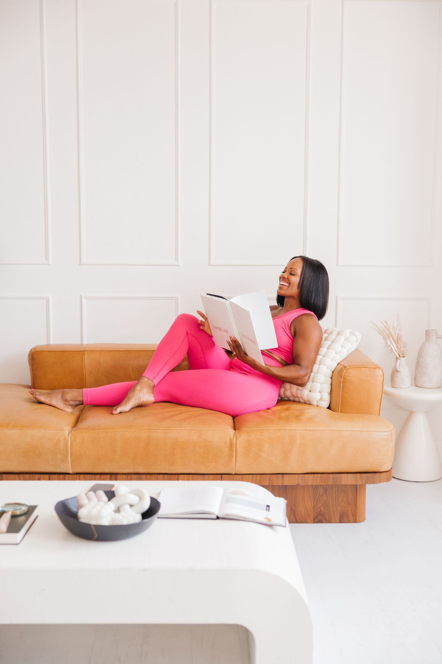 Smiling Black woman in bright pink outfit relaxing on tan leather sofa reading a book.