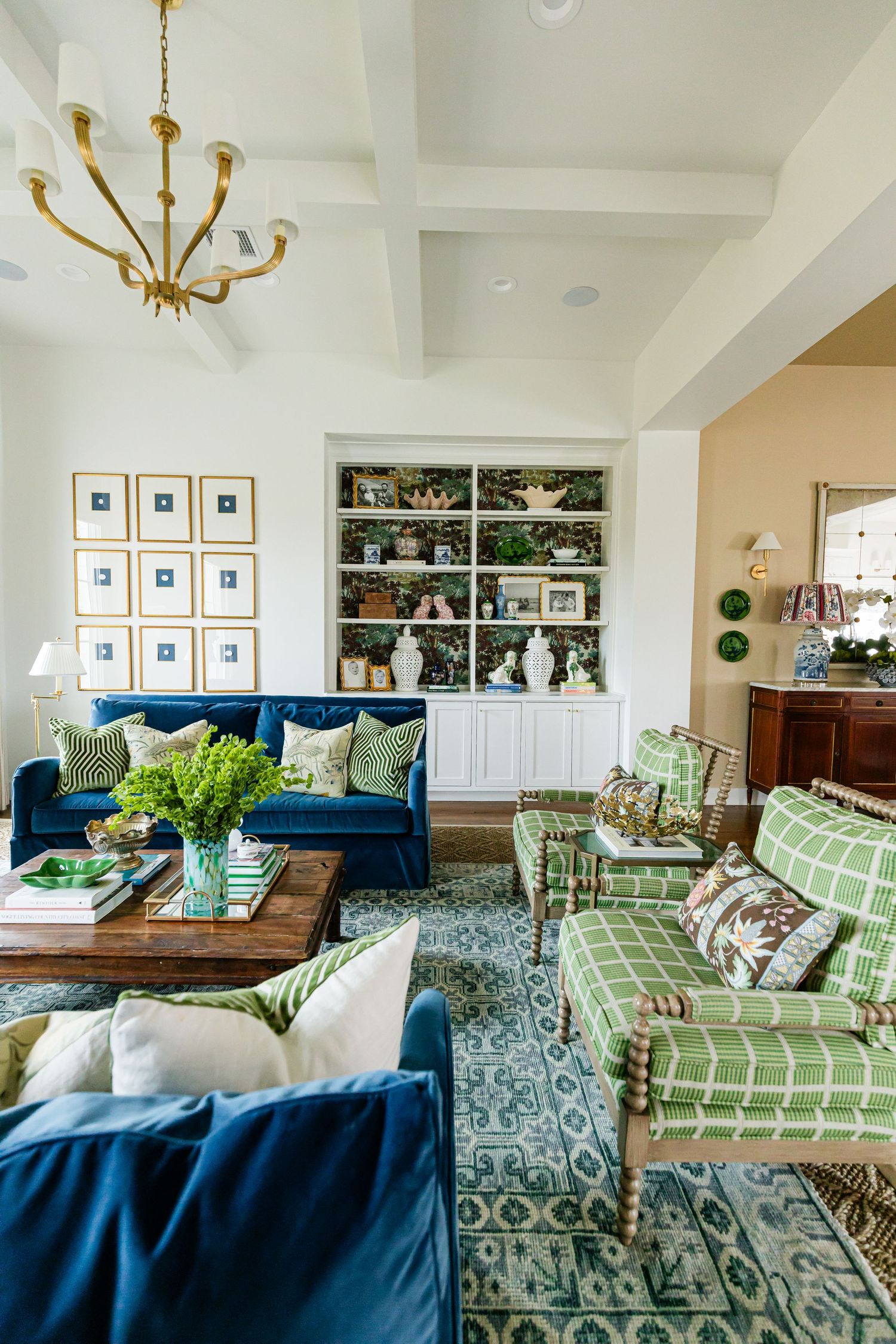 Elegant living room featuring navy velvet sofas, green plaid chairs, gold chandelier, and white built-in shelving.