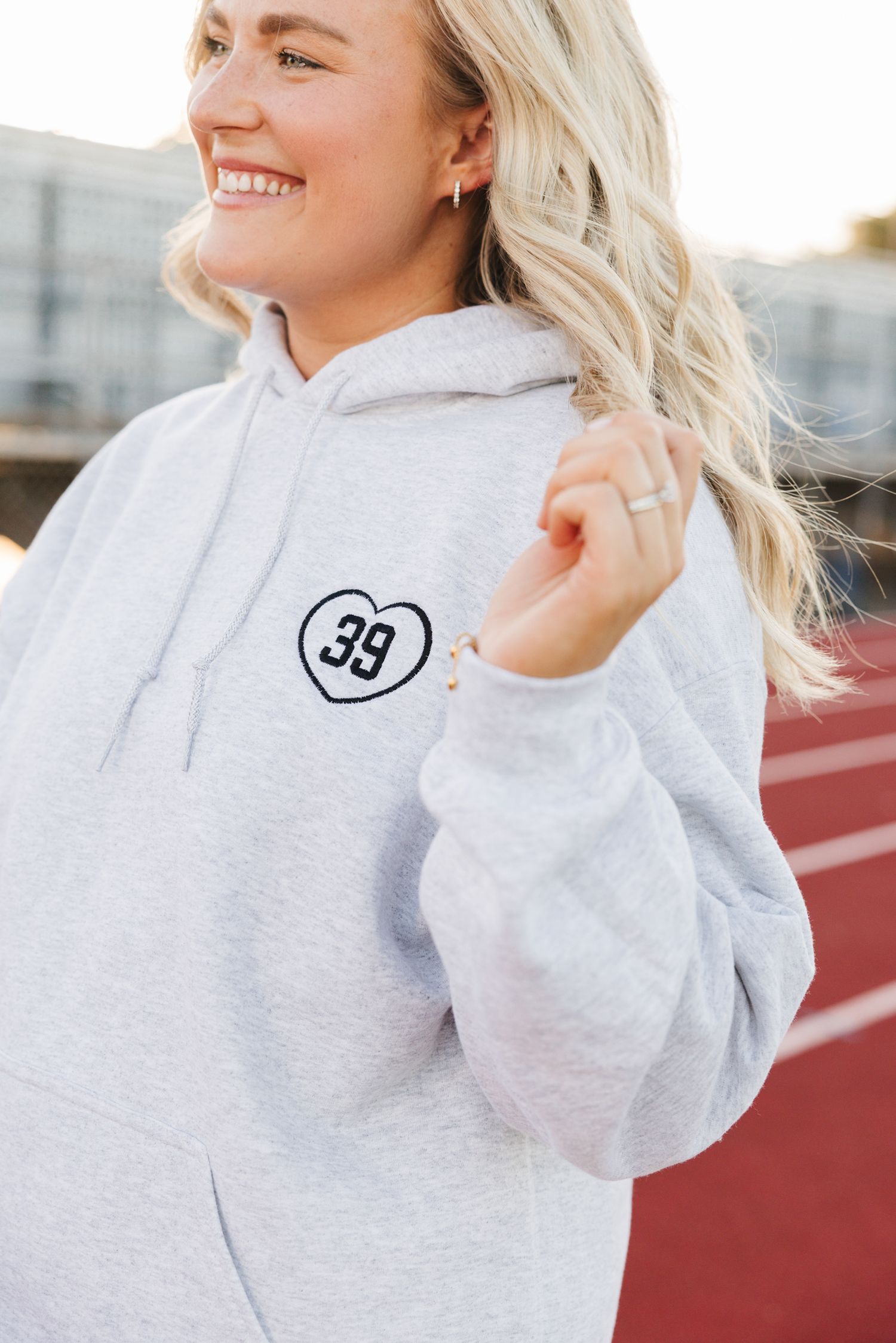 Smiling blonde woman wearing a gray hoodie with a heart number 39 embroidered patch on a track.