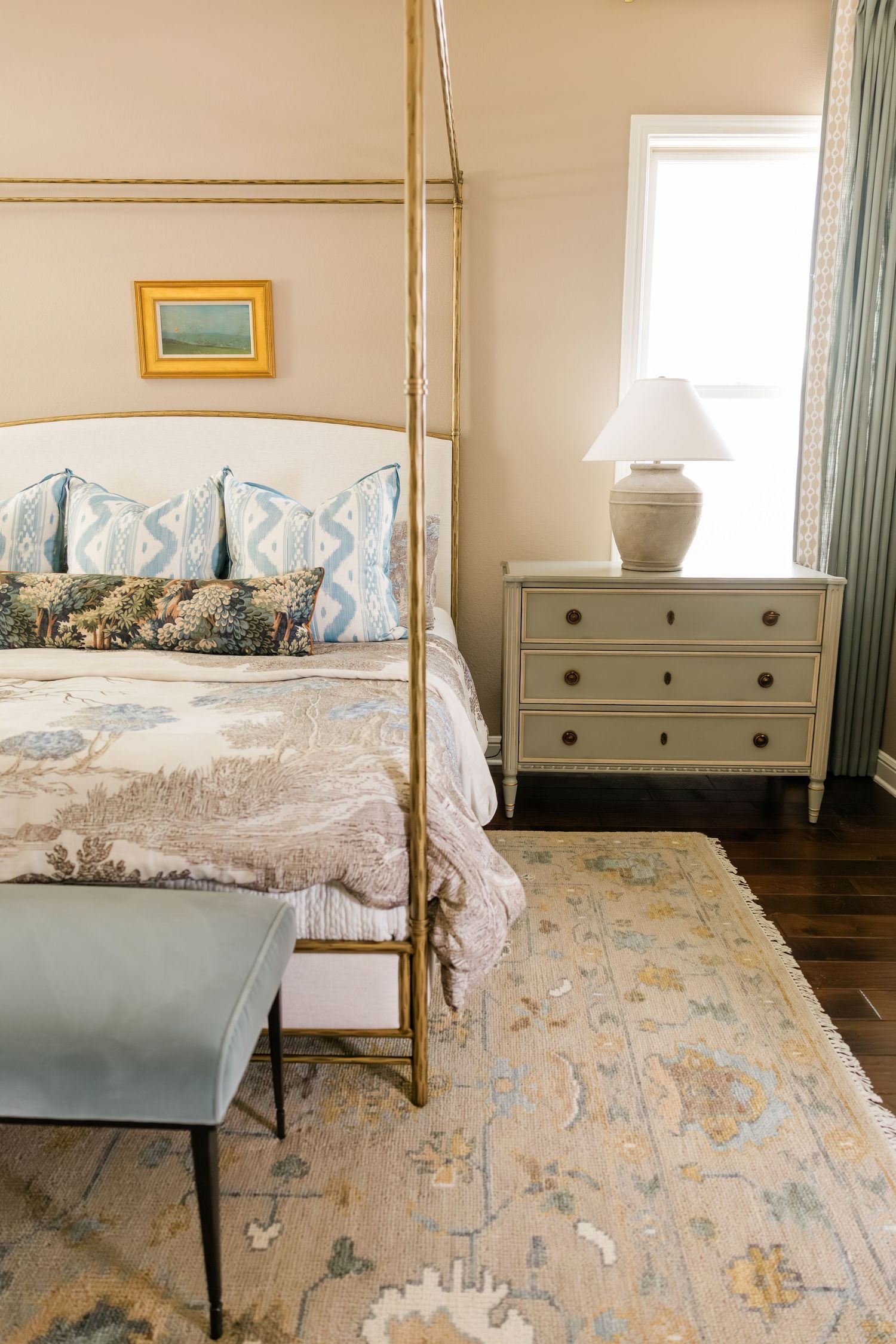 Elegant brass canopy bed with blue pillows, floral bedding, vintage rug, and white nightstand with lamp.