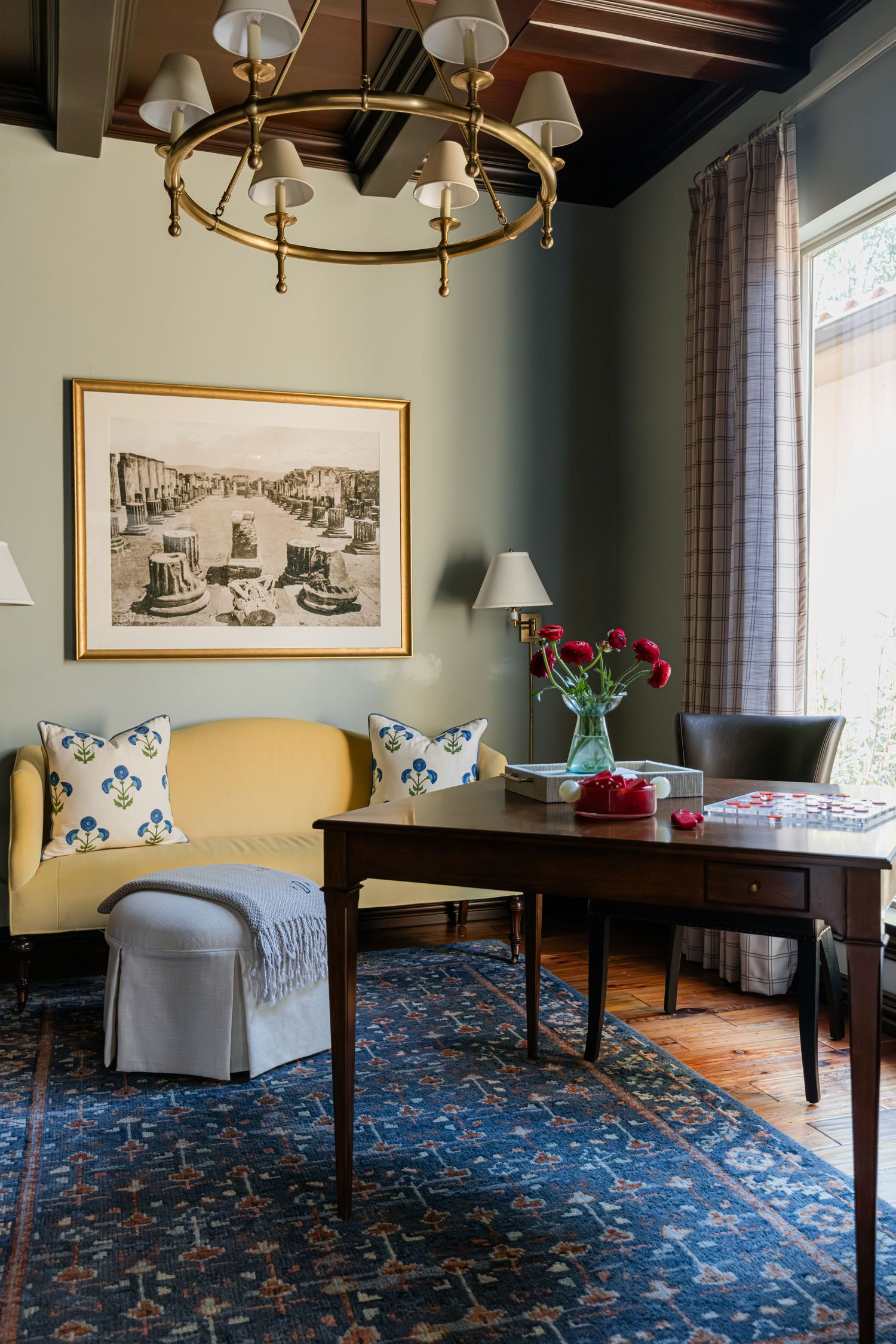 Elegant home office with yellow sofa, dark wood desk, brass chandelier, blue rug, and large framed artwork.
