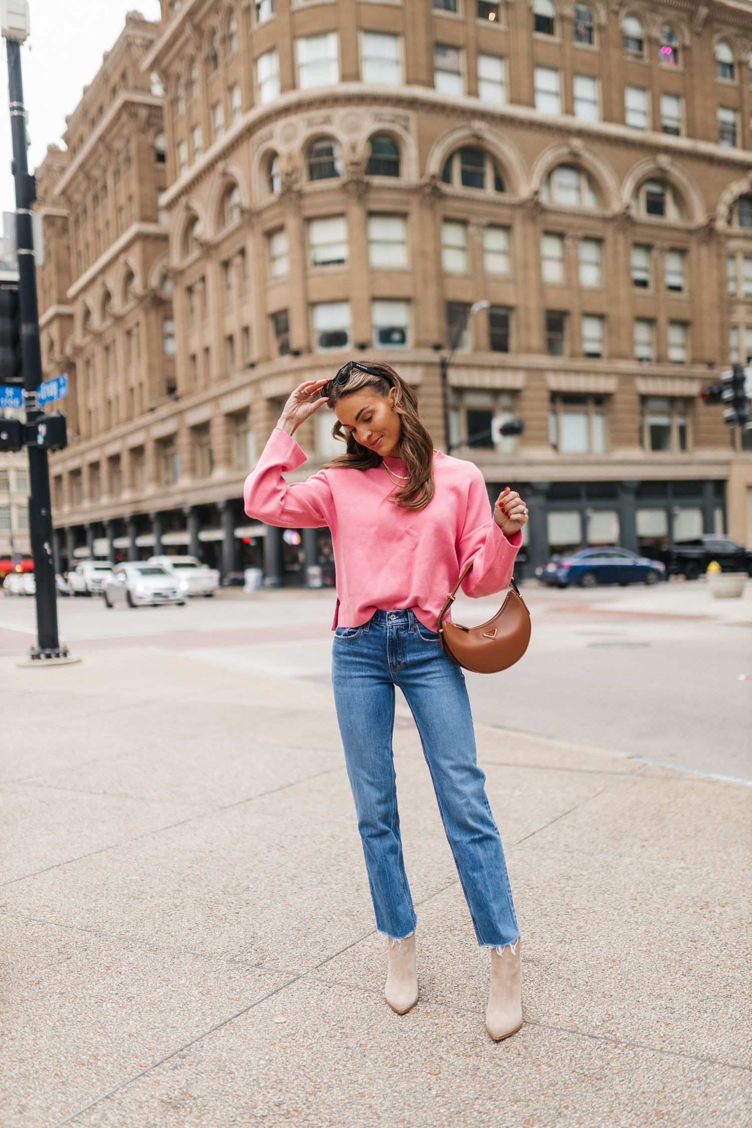 Woman in pink oversized sweater, straight-leg jeans, and cream ankle boots walking on a city street with brown bag.