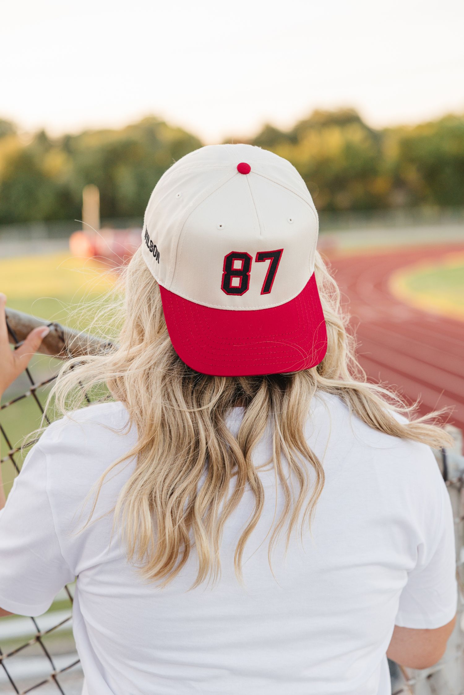 Back view of blonde woman wearing cream and red baseball cap with number 87 at a sports track.