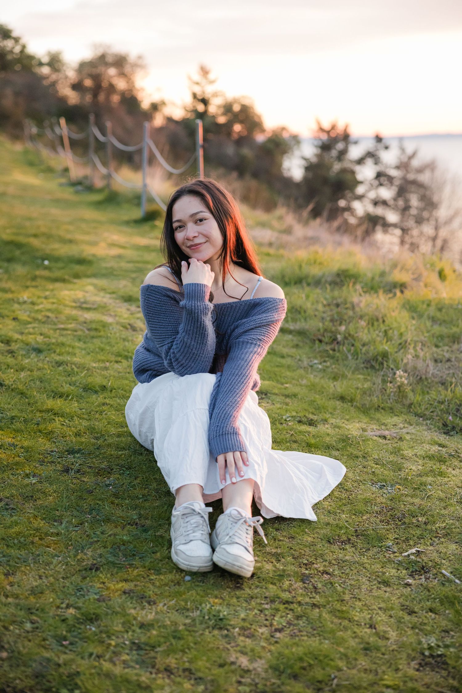 Young woman in gray off-shoulder sweater and white skirt sits relaxed on grassy hillside at sunset.