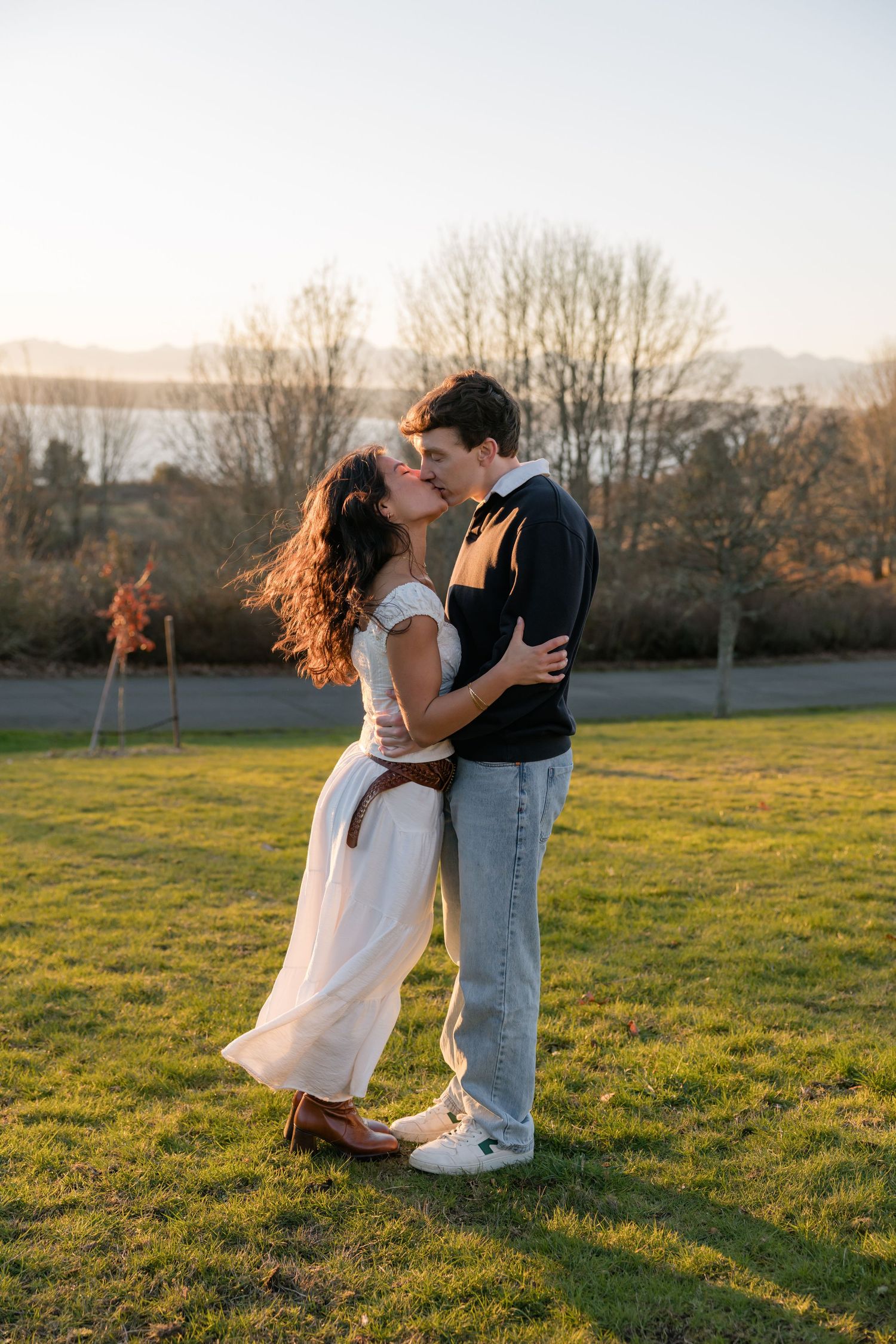 A couple kisses at golden hour in a grassy park with mountains and a lake in the distance.