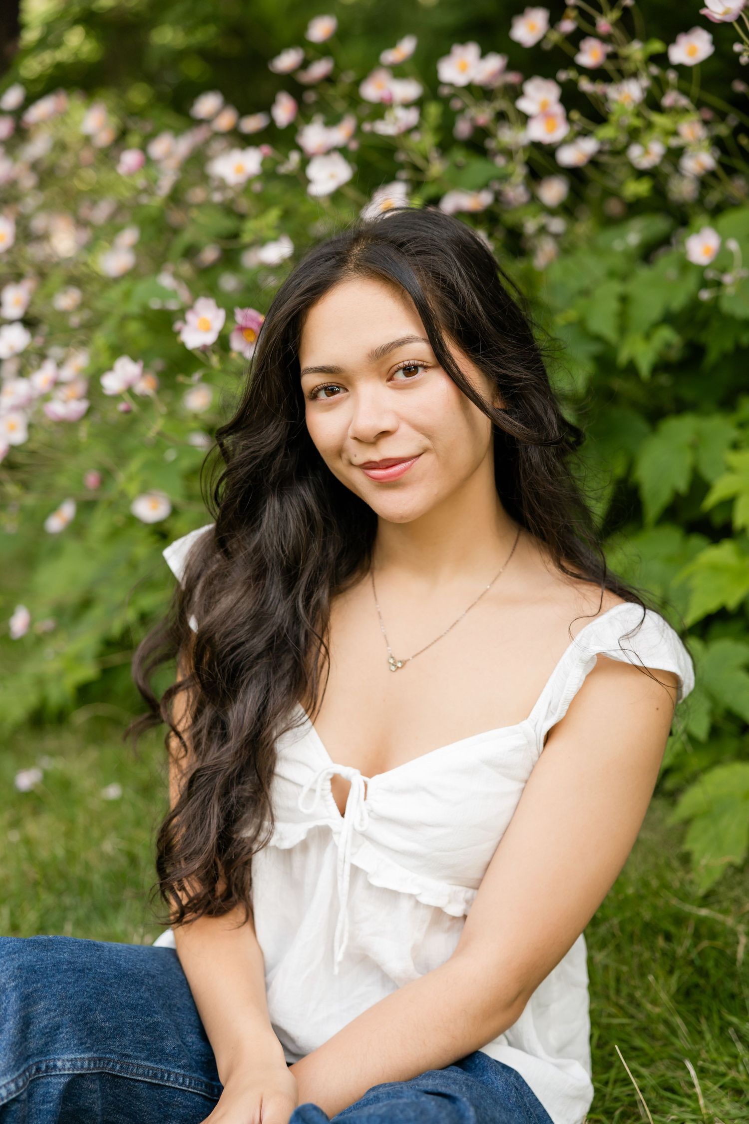 Young woman with long dark wavy hair in a white top and jeans sits in front of blooming pink flowers.