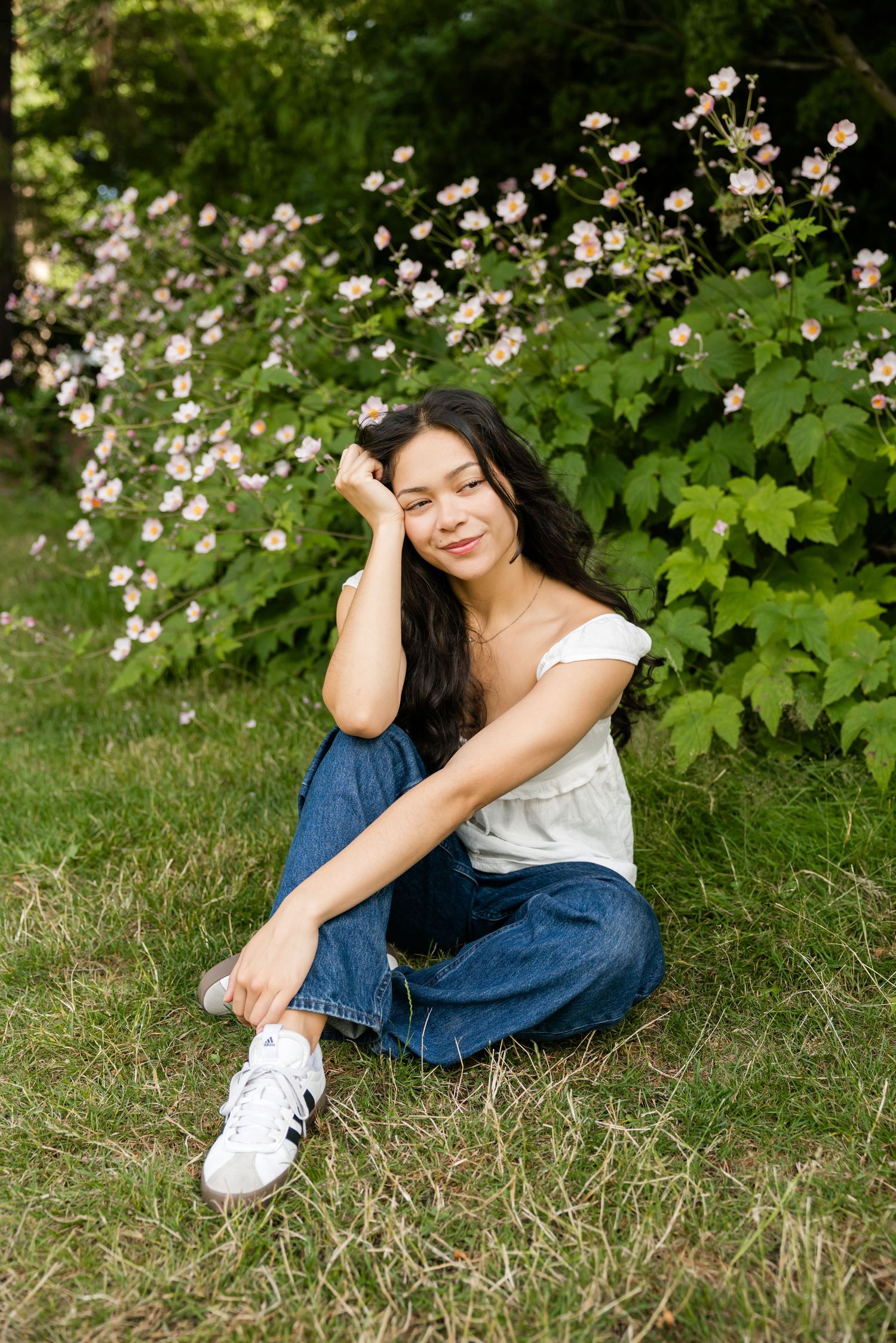 Young woman with eyes closed sits peacefully on grass beside lush pink flowering bushes in a garden.