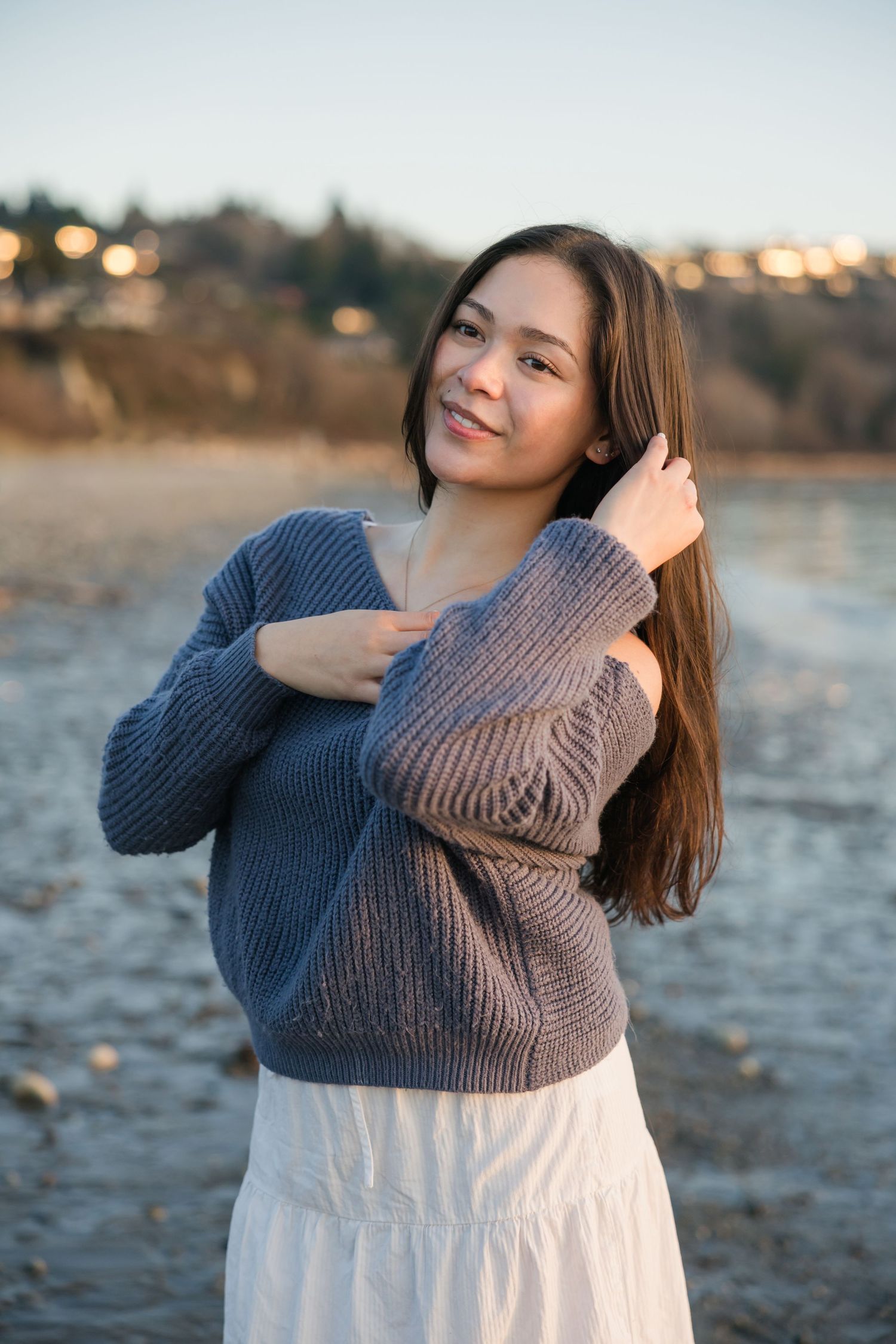 Young woman in gray knit sweater and white skirt poses gracefully on a rocky beach at golden hour.