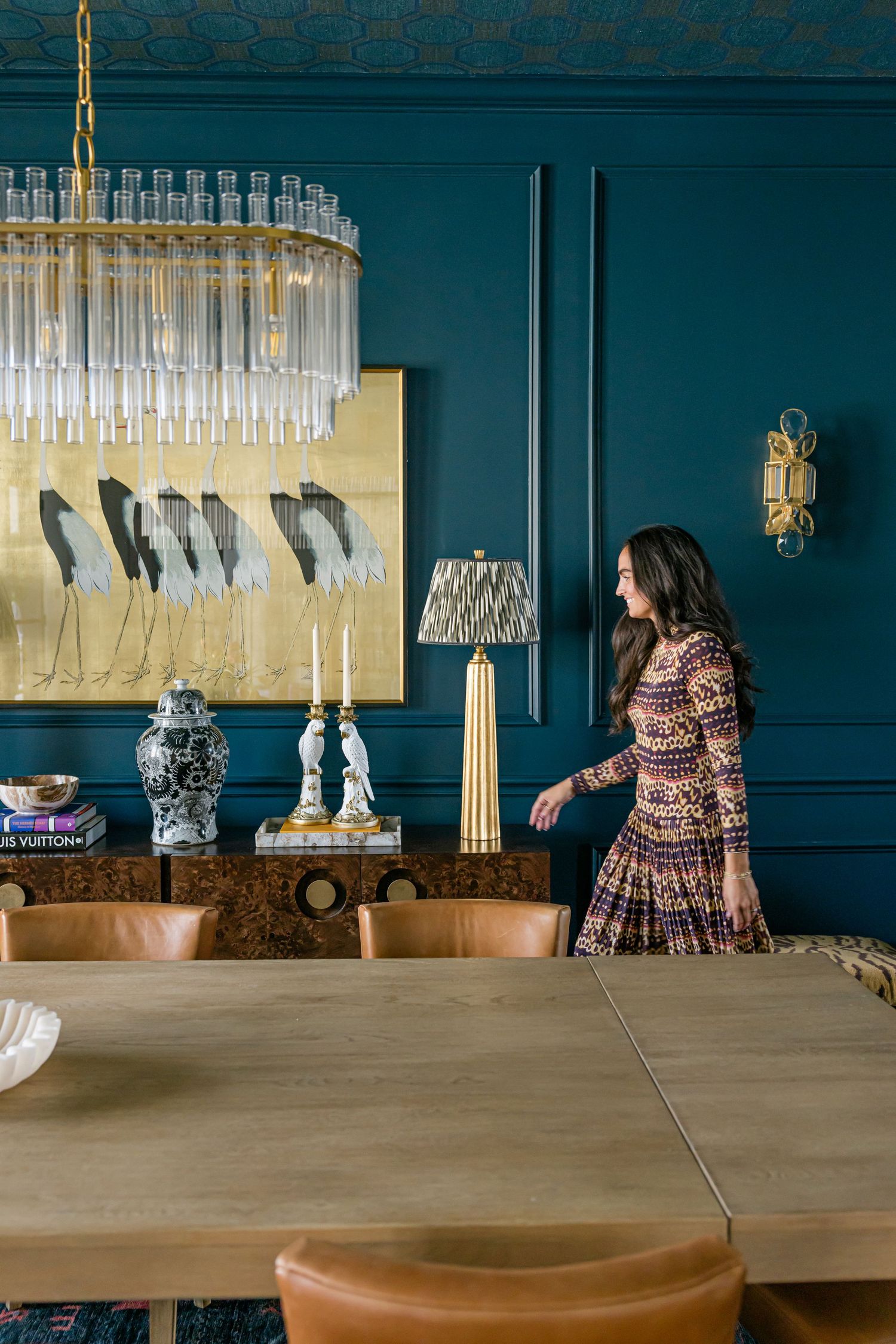 Woman in patterned dress in teal dining room with crystal chandelier, gold artwork, and burl wood sideboard.