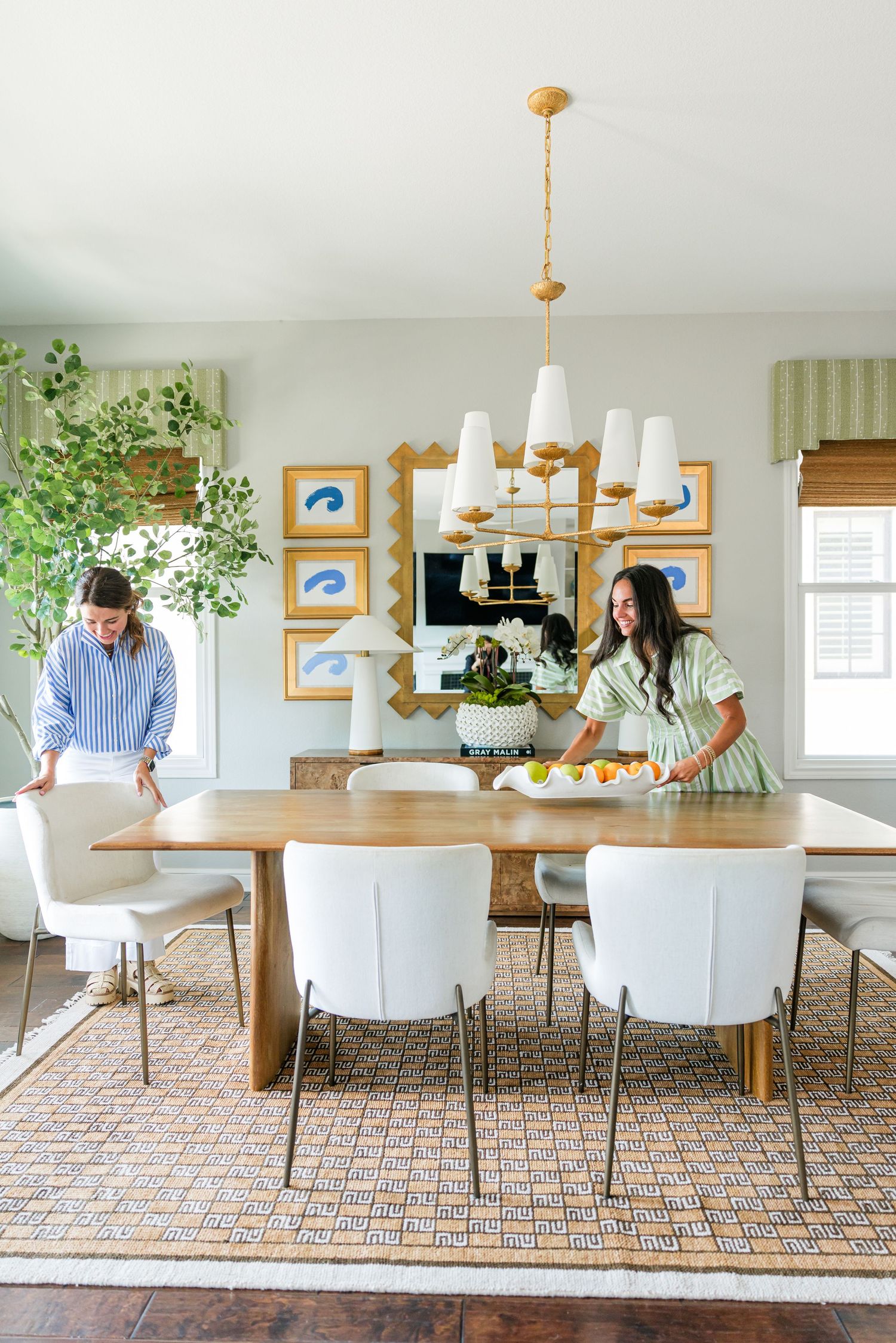 Two women arranging a wood dining table with white chairs beneath a gold chandelier in a light-filled room.