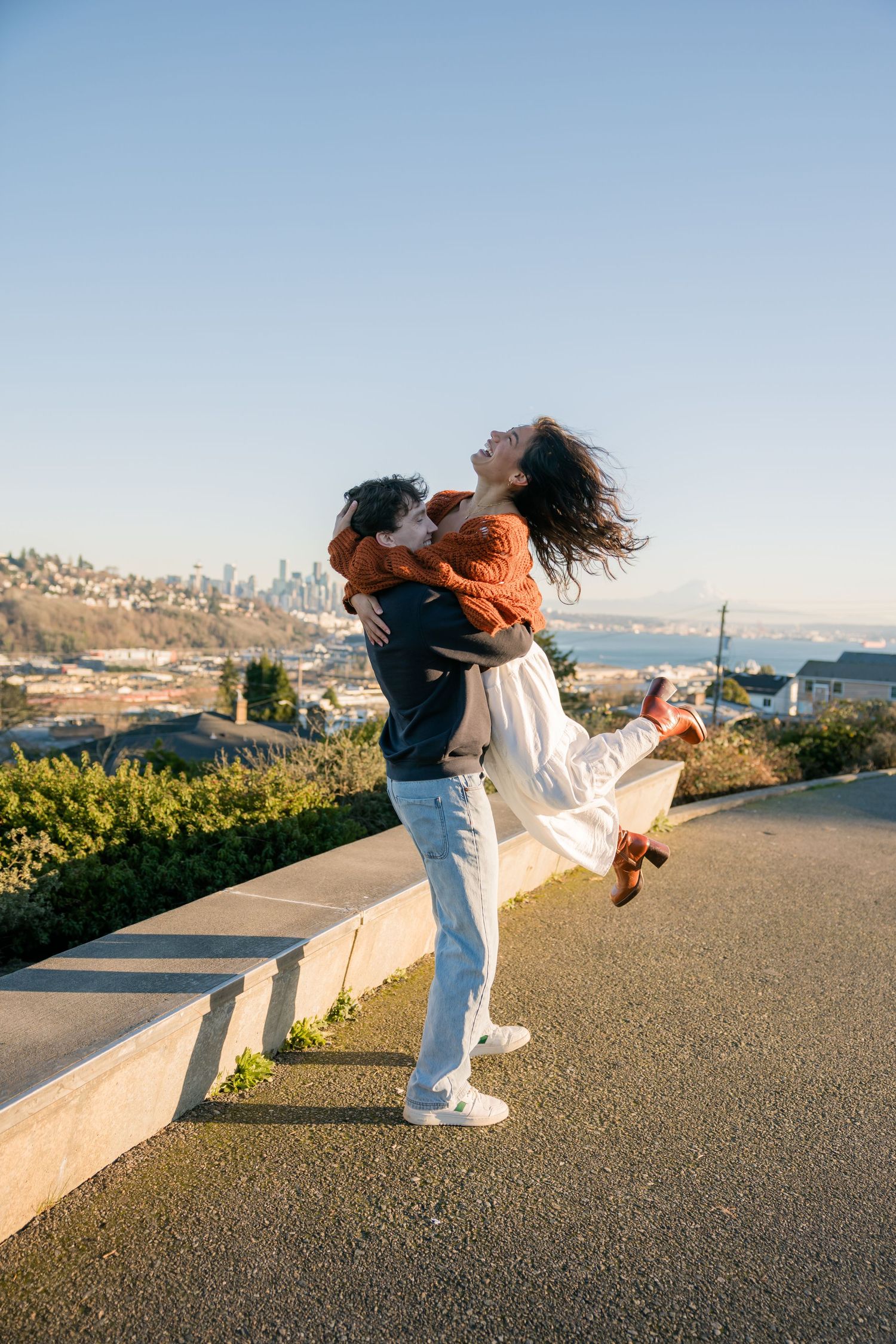 A man joyfully lifts a woman in a white dress with a city skyline and bay behind them.
