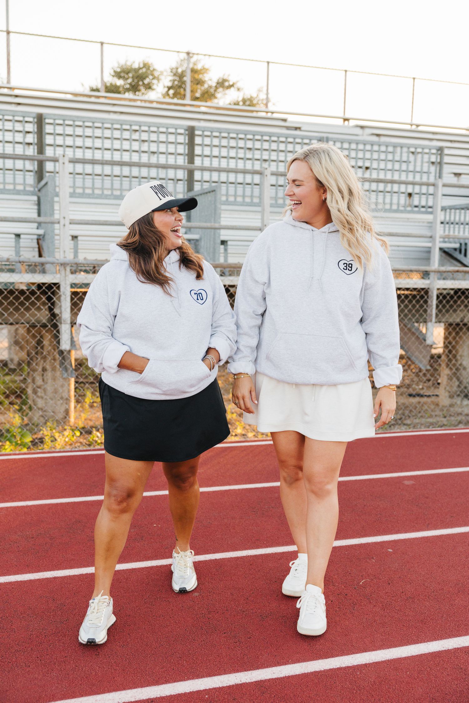 Two women in matching gray hoodies with number patches walking and laughing on a running track.