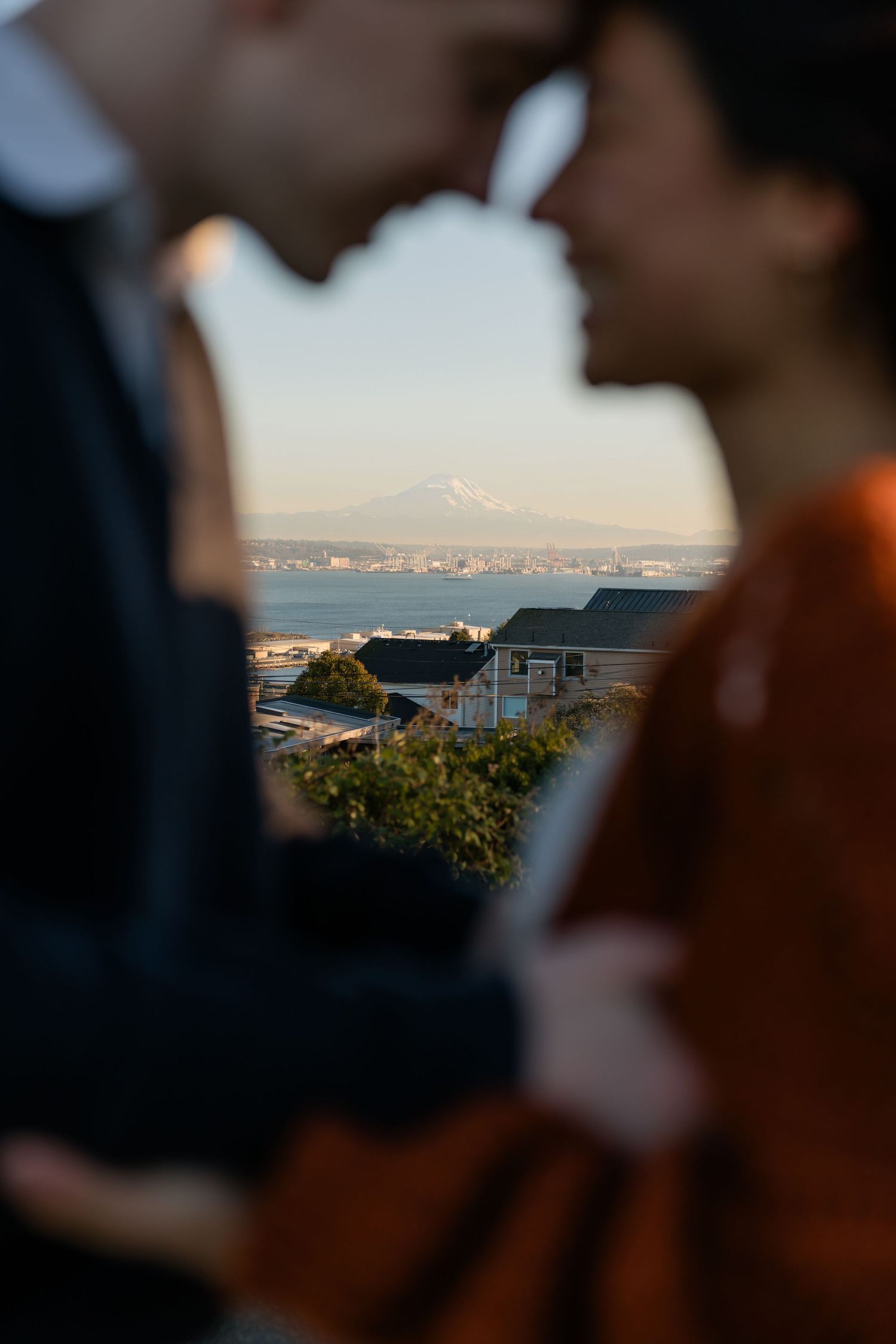 A couple shares a romantic moment with a snow-capped mountain and bay visible in the background.