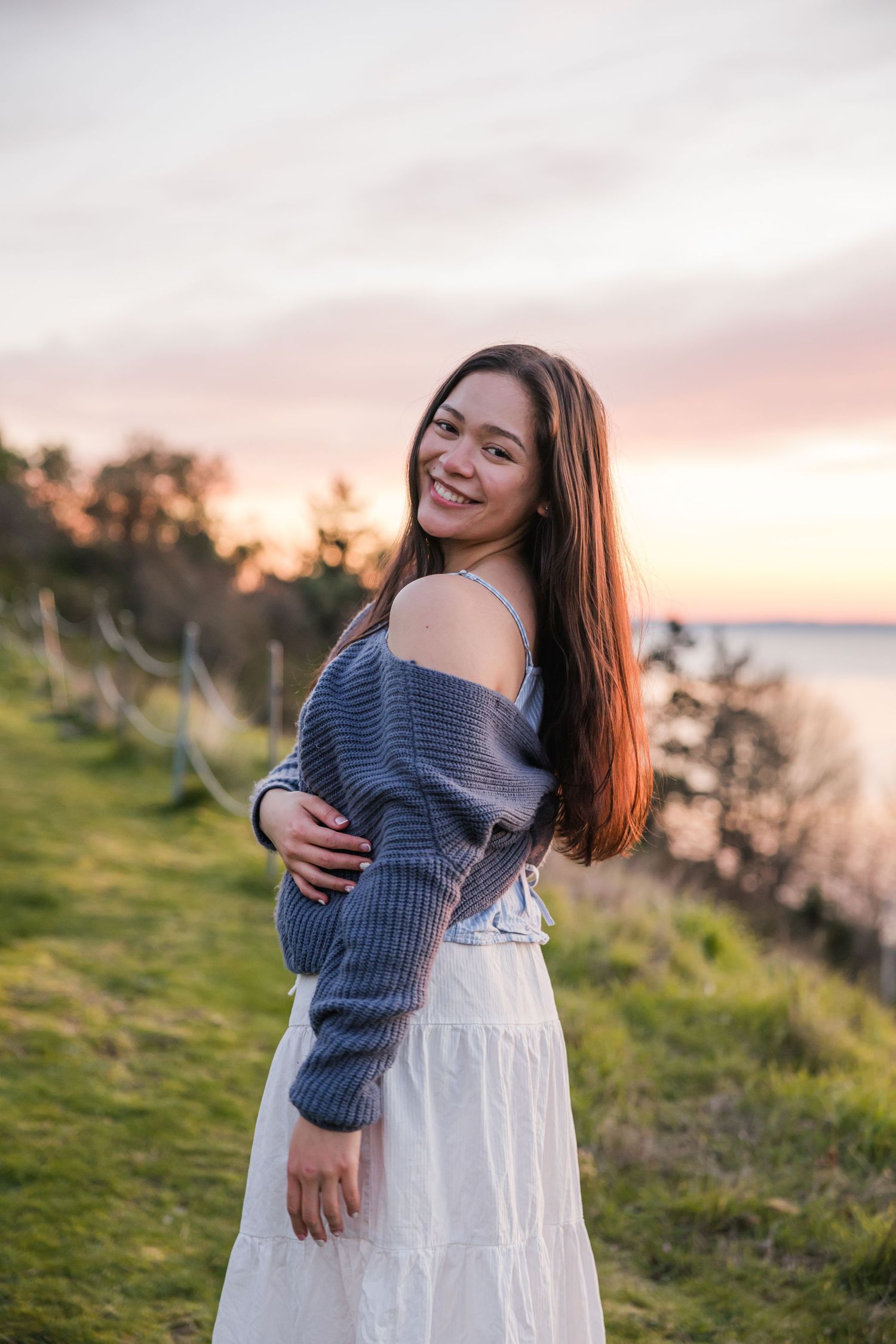 Smiling young woman in off-shoulder gray sweater and white skirt poses on grassy cliff at sunset.