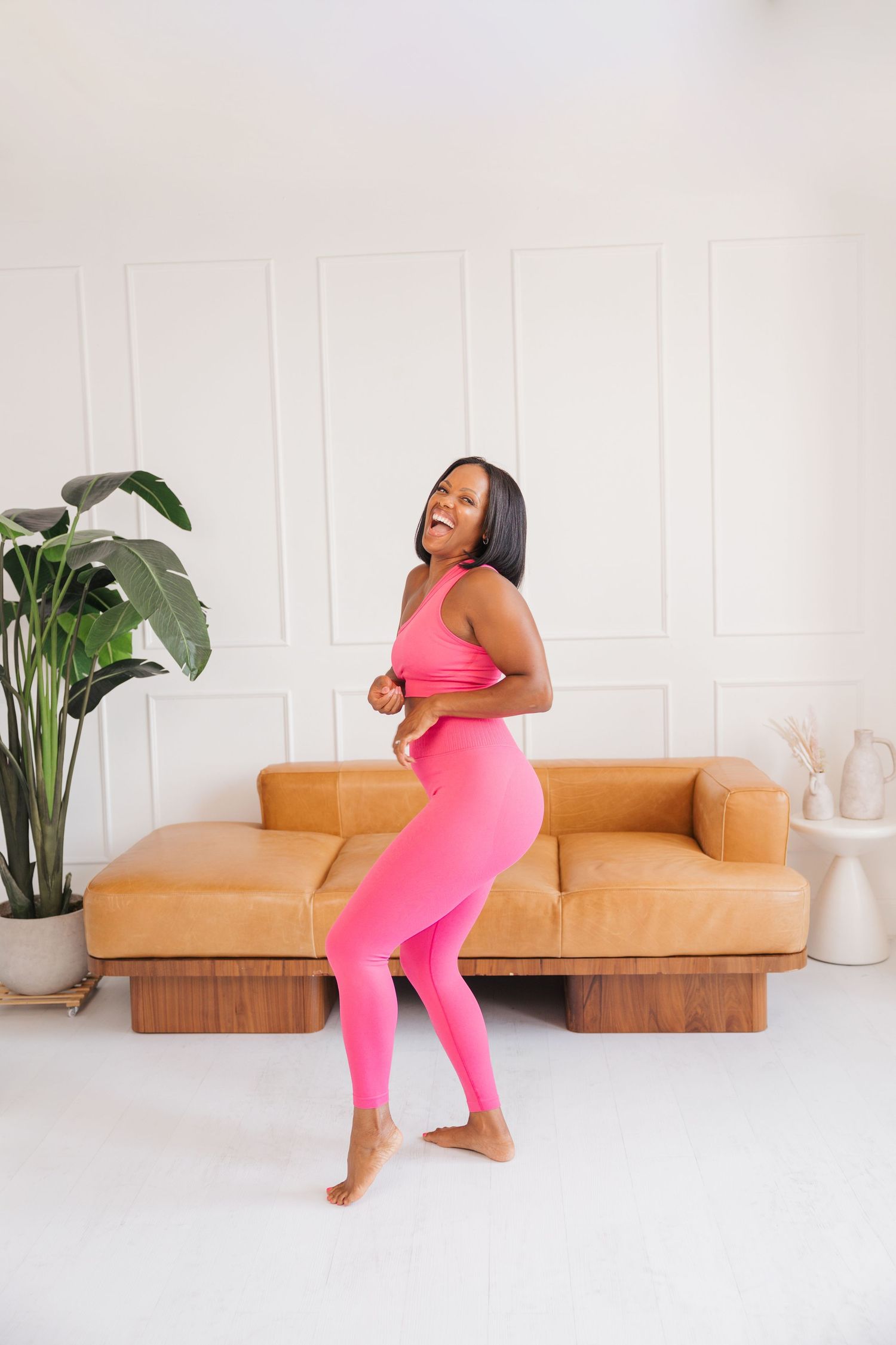 Joyful Black woman in hot pink athletic set dancing barefoot in bright modern living room.