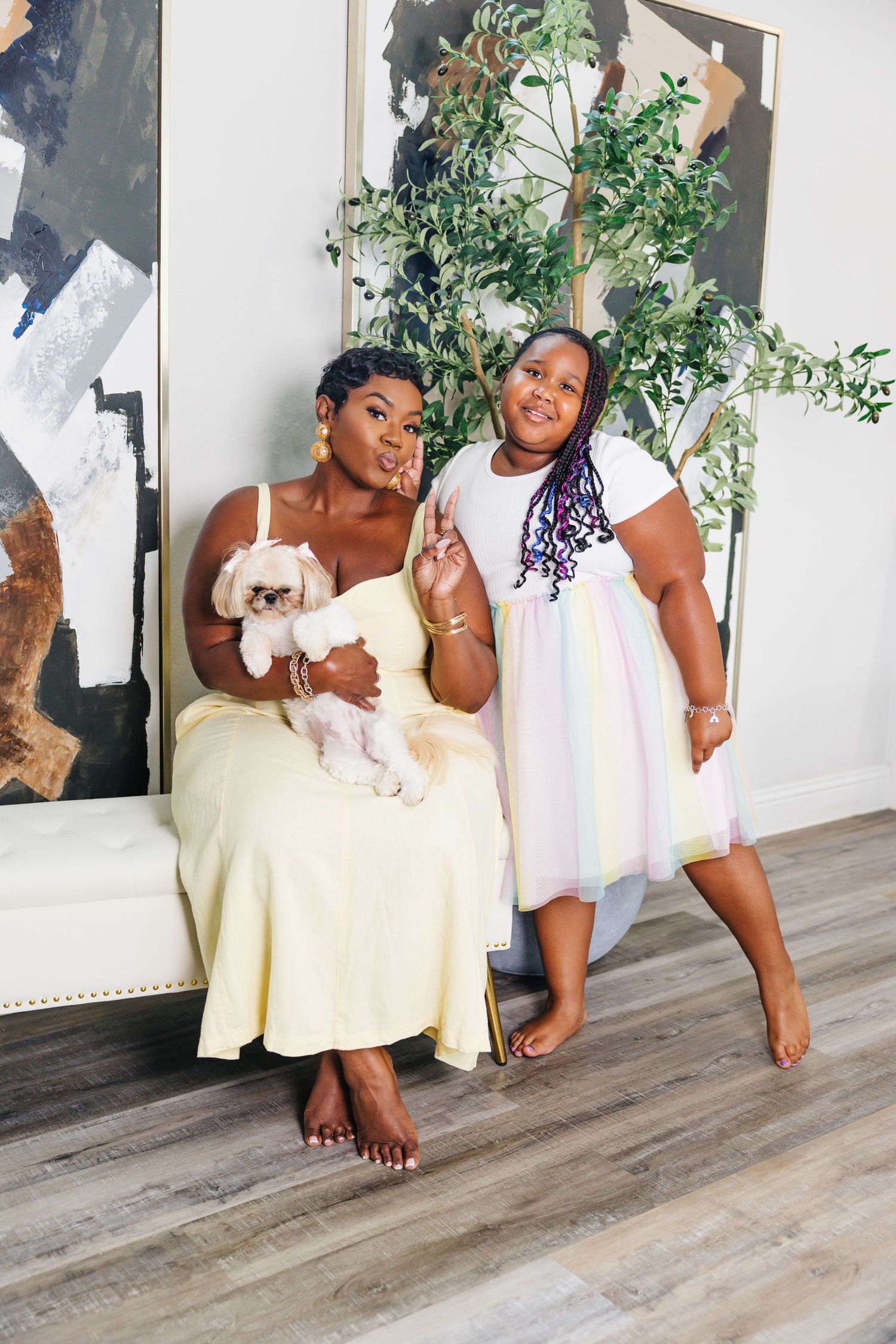 A smiling woman in a yellow dress holding a small dog poses with a young girl in a rainbow dress indoors.
