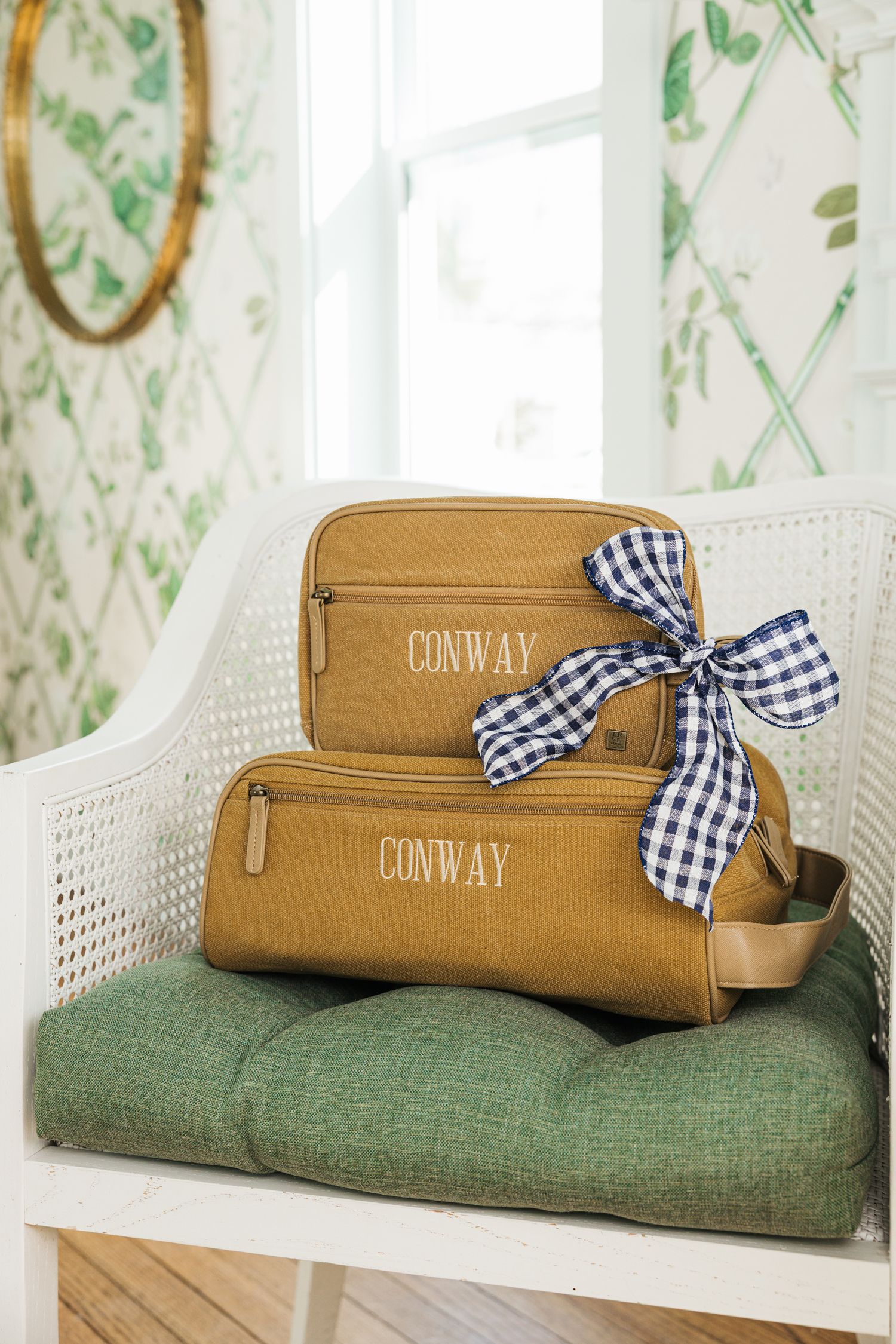 Two tan canvas monogrammed Conway toiletry bags stacked on white chair with navy gingham bow.