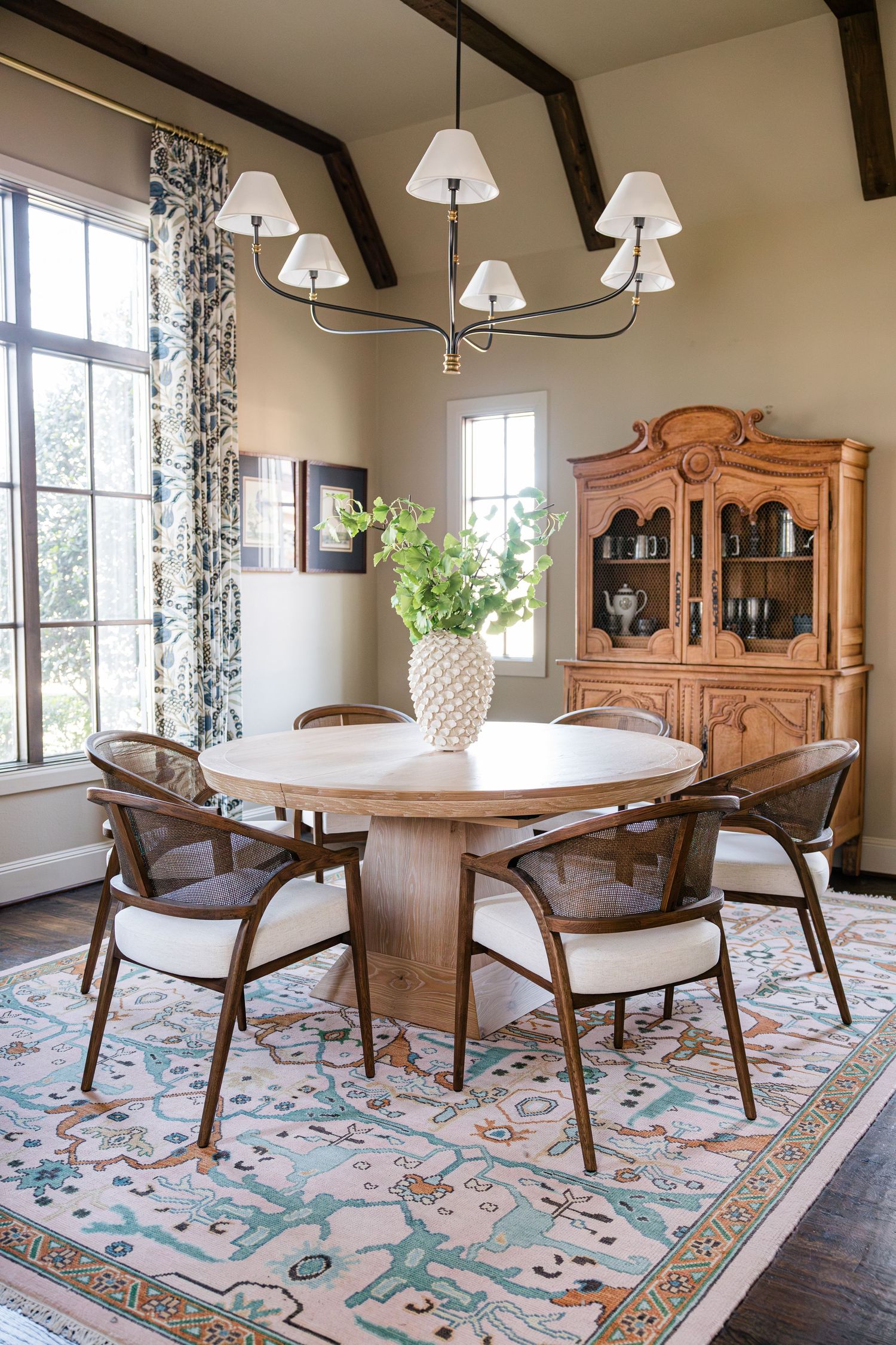 Round wood dining table with wicker chairs, white vase, and ornate hutch under a multi-shade chandelier.