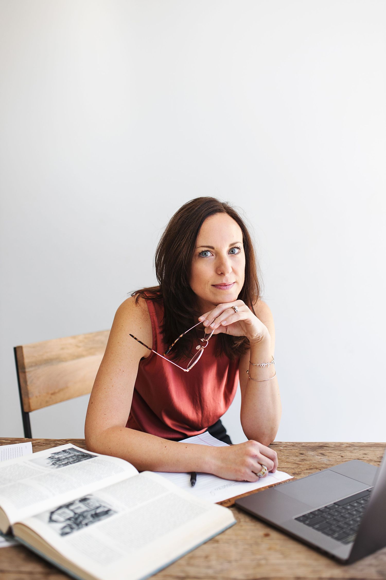 Woman in red top sitting at wooden desk with open book and laptop, holding glasses thoughtfully.