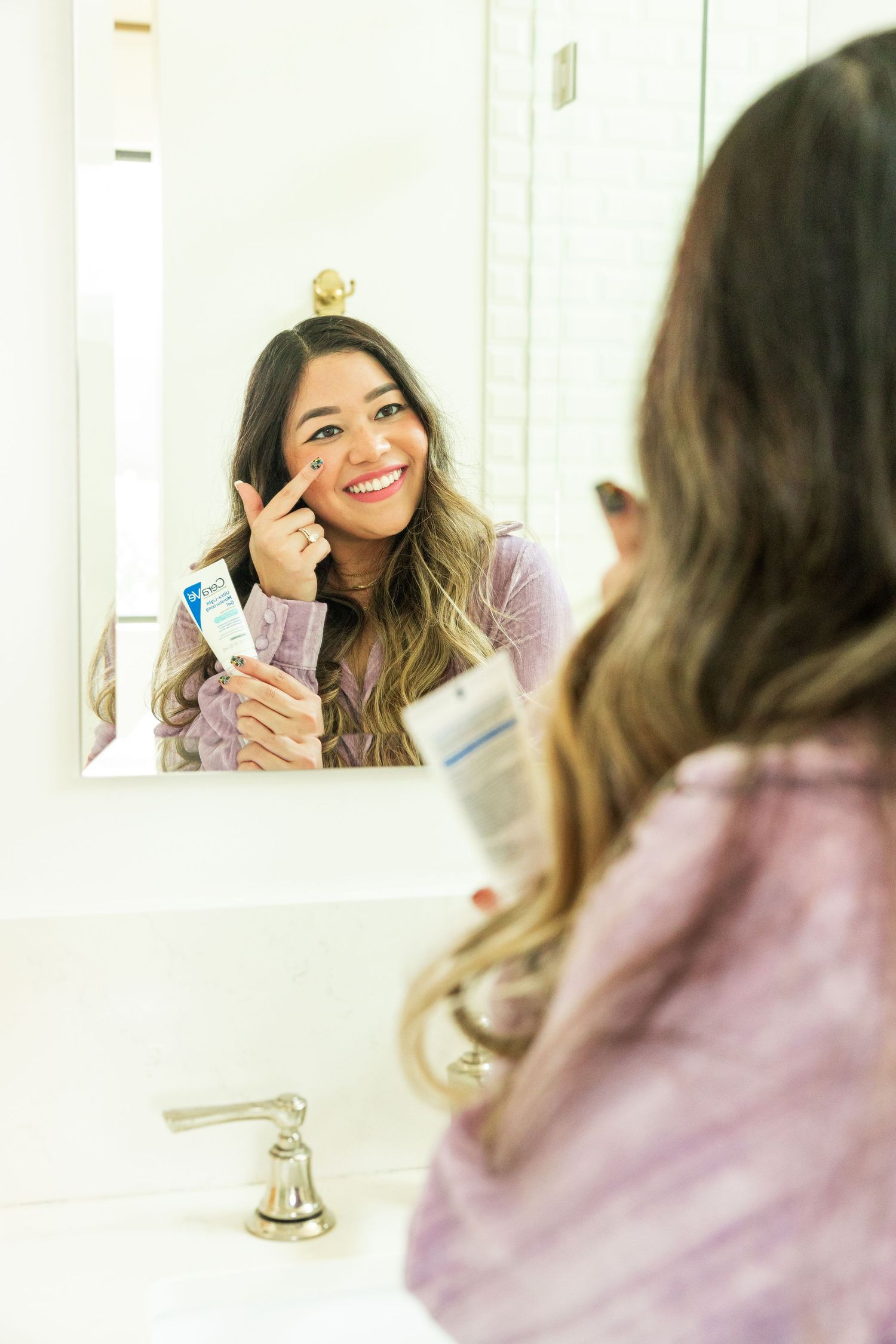 Smiling woman applying skincare product while looking in a bathroom mirror wearing a purple robe.