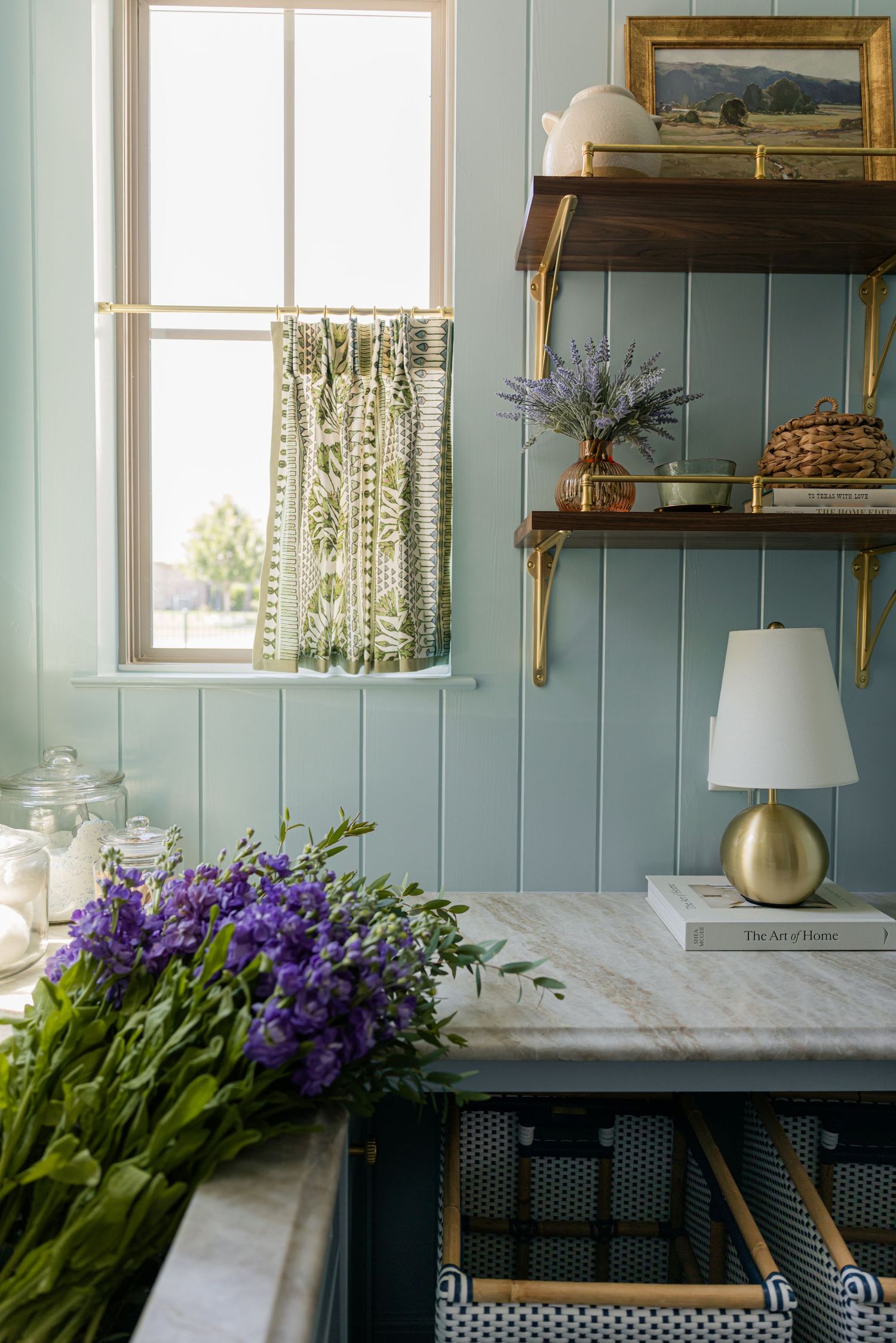 Blue laundry room detail with wood shelves, brass brackets, gold lamp, purple flowers, and patterned cafe curtain.