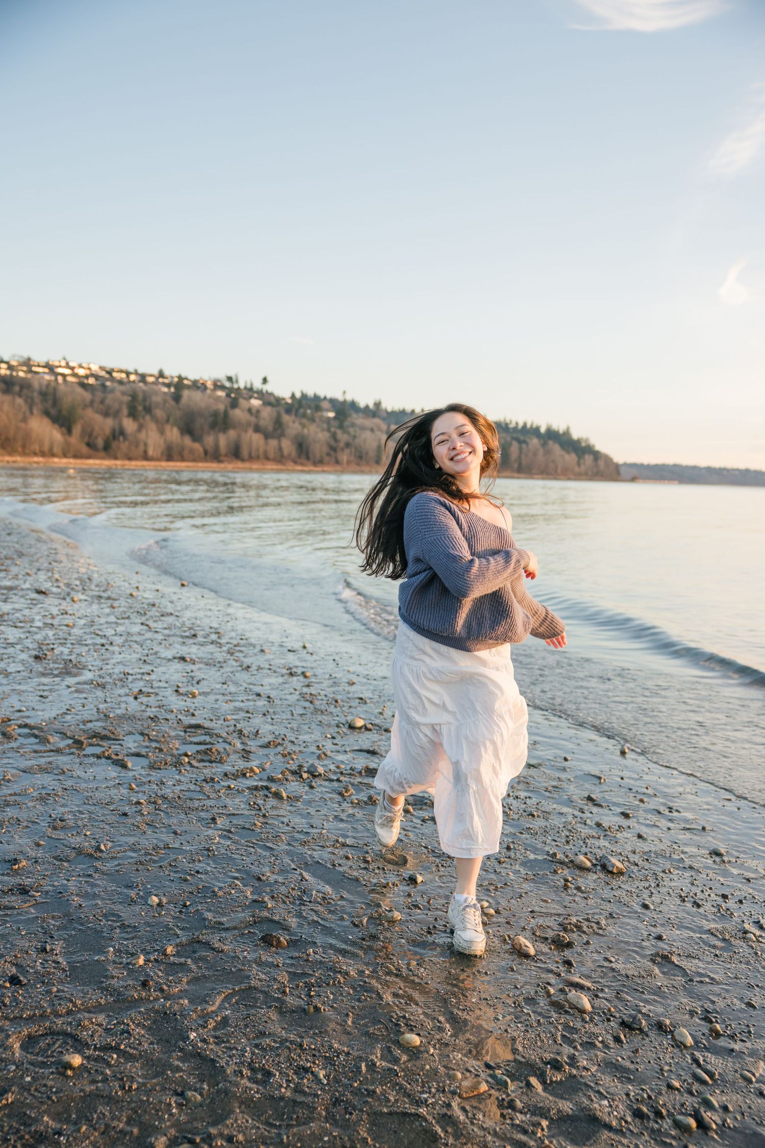 Joyful young woman in gray sweater and white skirt running along a sandy beach shoreline at sunset.